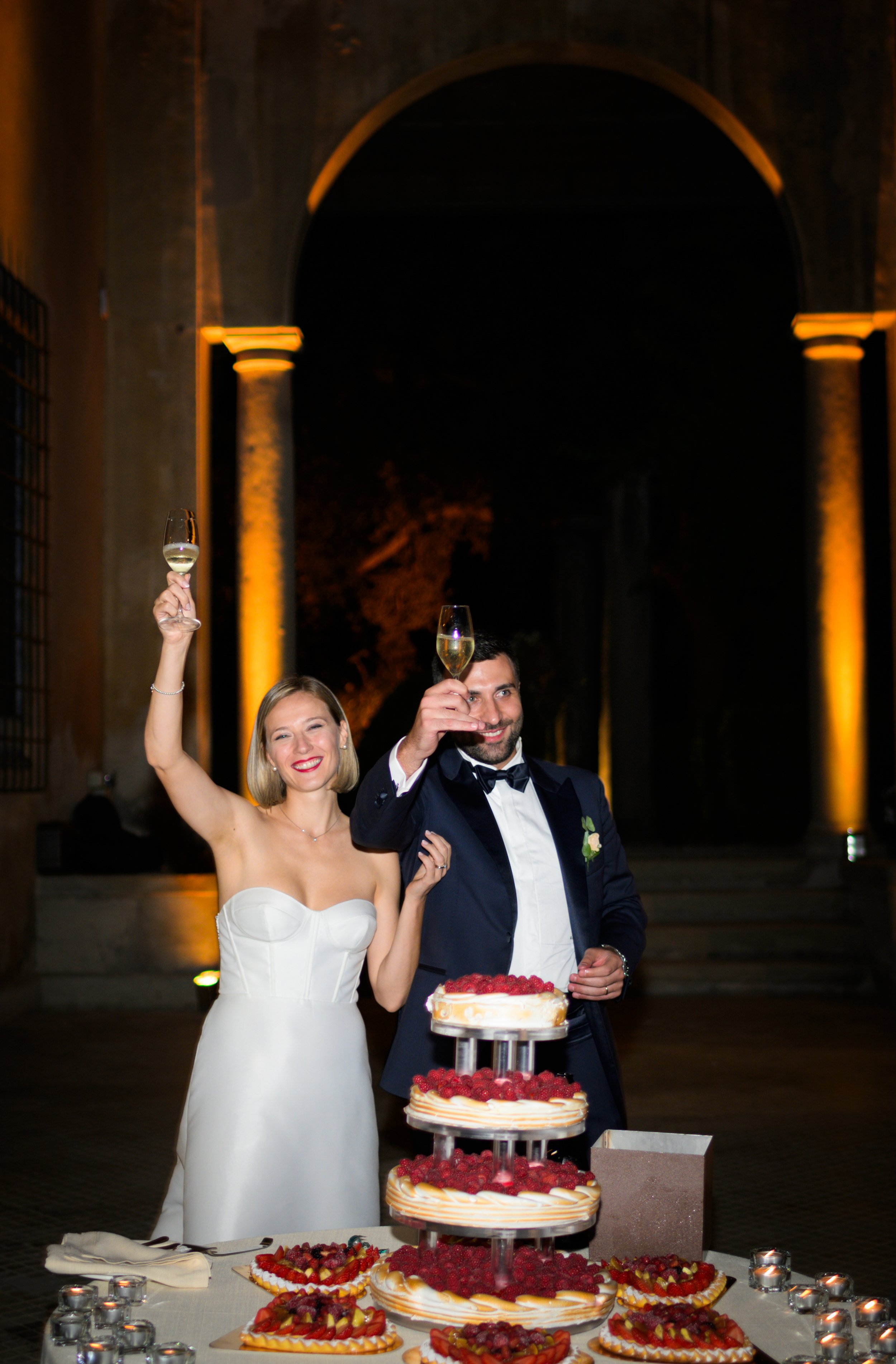 Una coppia felice in abito da cerimonia brinda con bicchieri di spumante davanti a una torre di torte decorate con frutta. L'ambiente è elegante e ben illuminato.