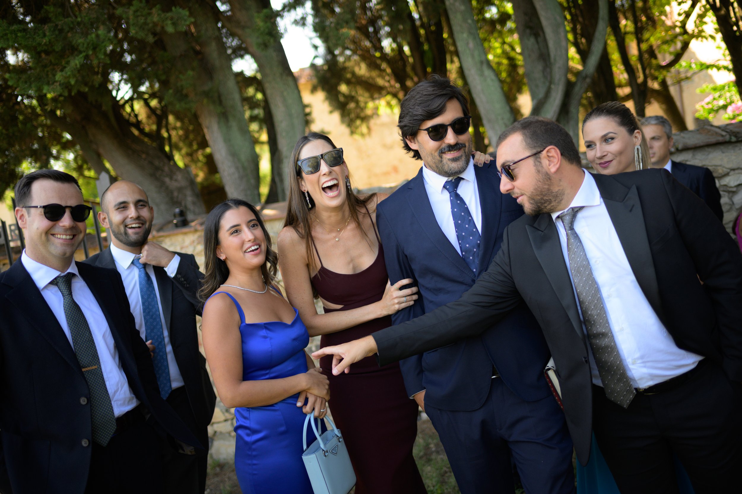 Gruppo di persone eleganti che ridono all'aperto, indossano abiti formali.