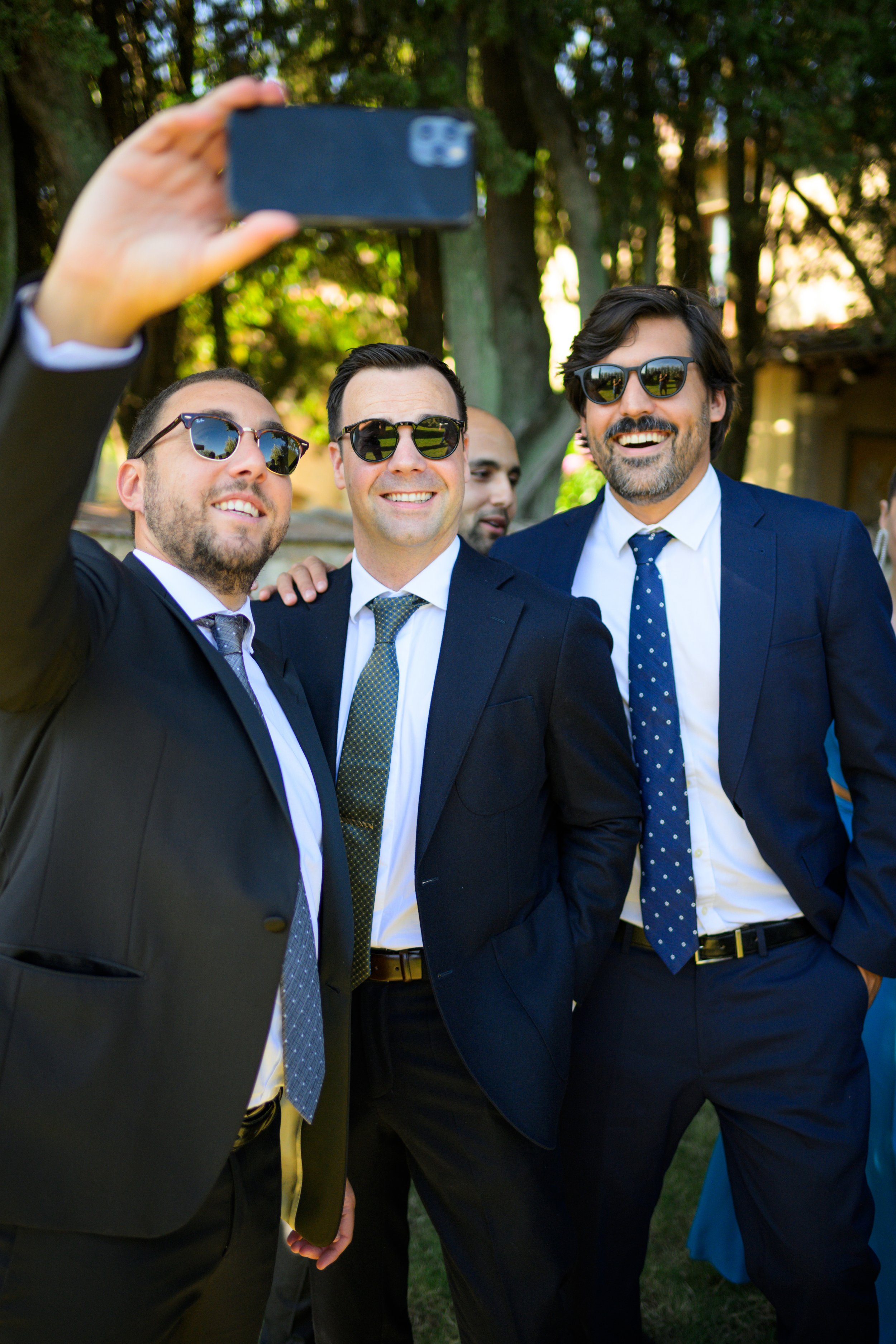 Quattro uomini in abiti eleganti scattano un selfie insieme all'aperto, sorridenti e con occhiali da sole.