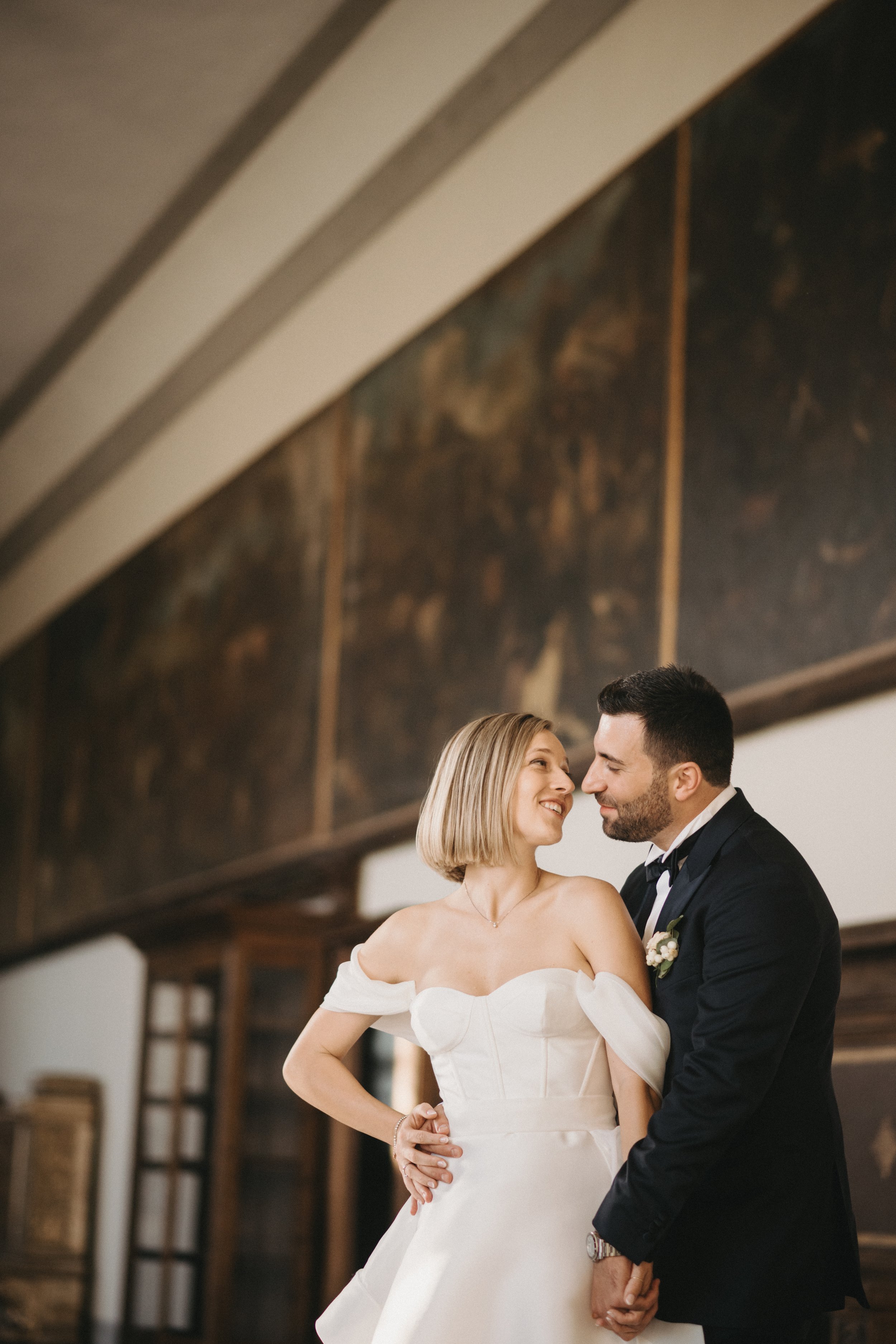 Una coppia vestita elegantemente, con la sposa in abito bianco, sorride e si tiene per mano in un ambiente interno con decorazioni classiche.