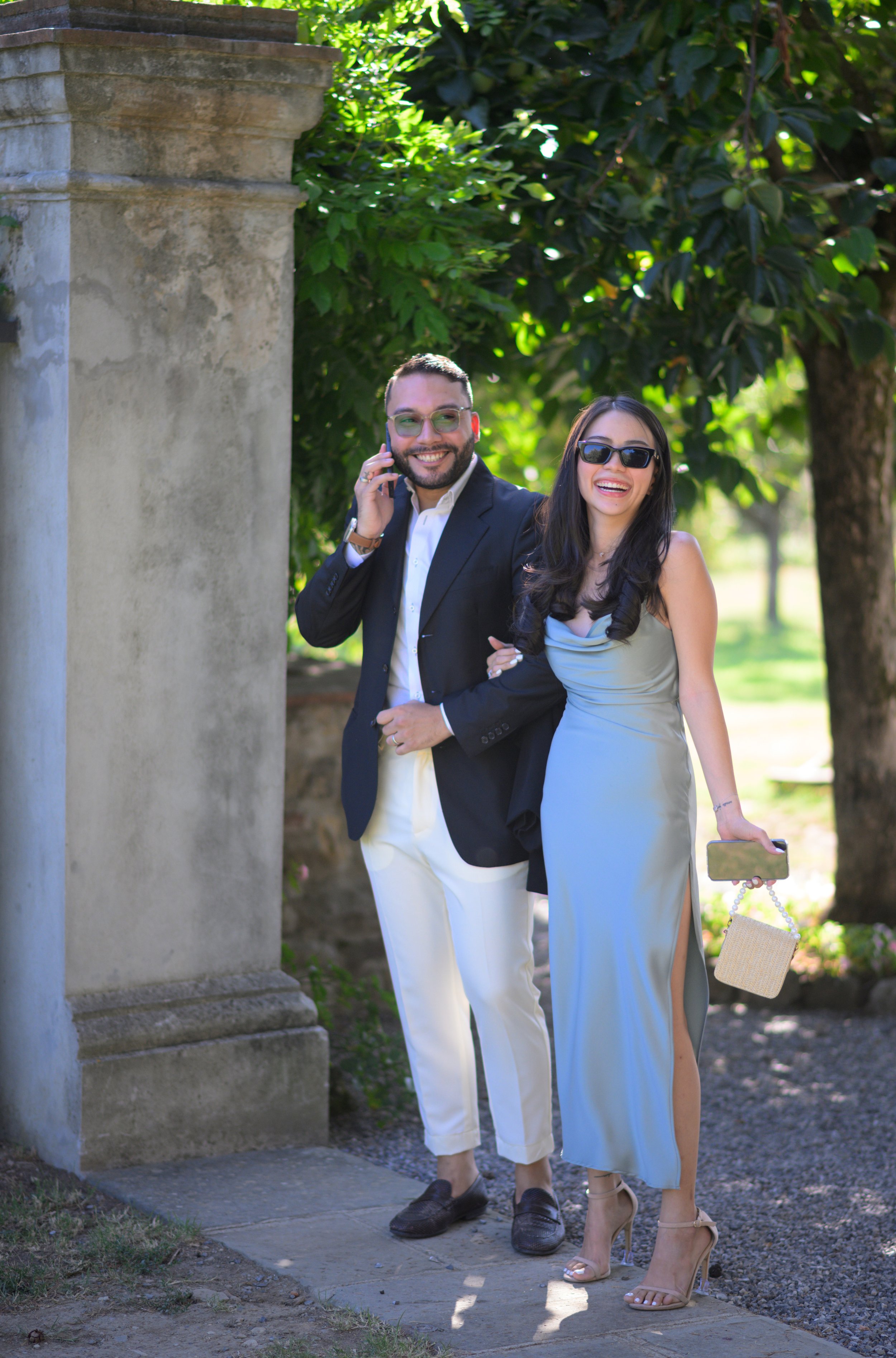 Due persone sorridenti in piedi in un giardino, una con abito lungo azzurro e l'altra in completo giacca scura e pantaloni chiari. La donna tiene una piccola borsetta e un cellulare, mentre l'uomo parla al telefono.