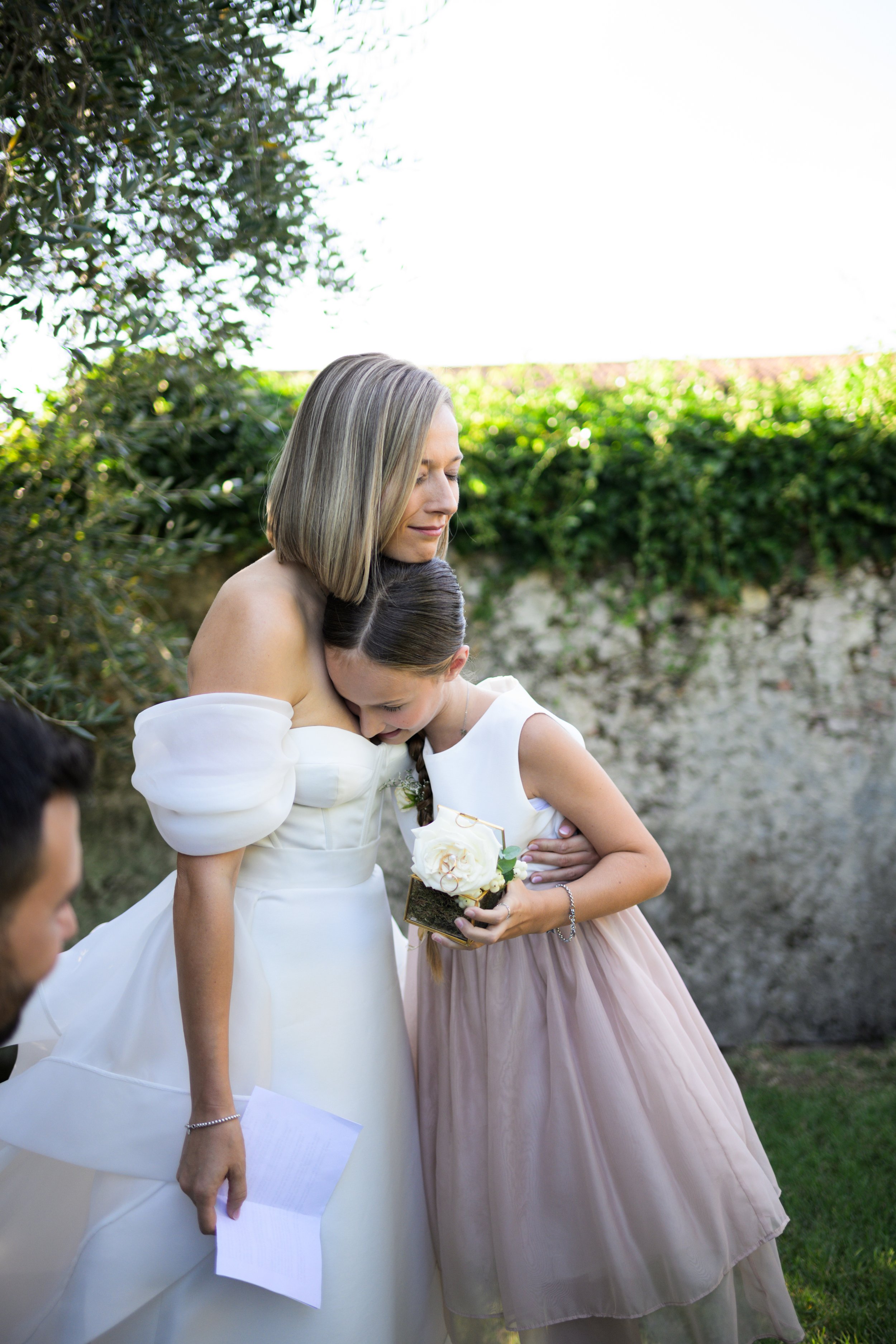 Donna in abito da sposa abbraccia una bambina con un abito elegante e una rosa bianca in mano, all'aperto.
