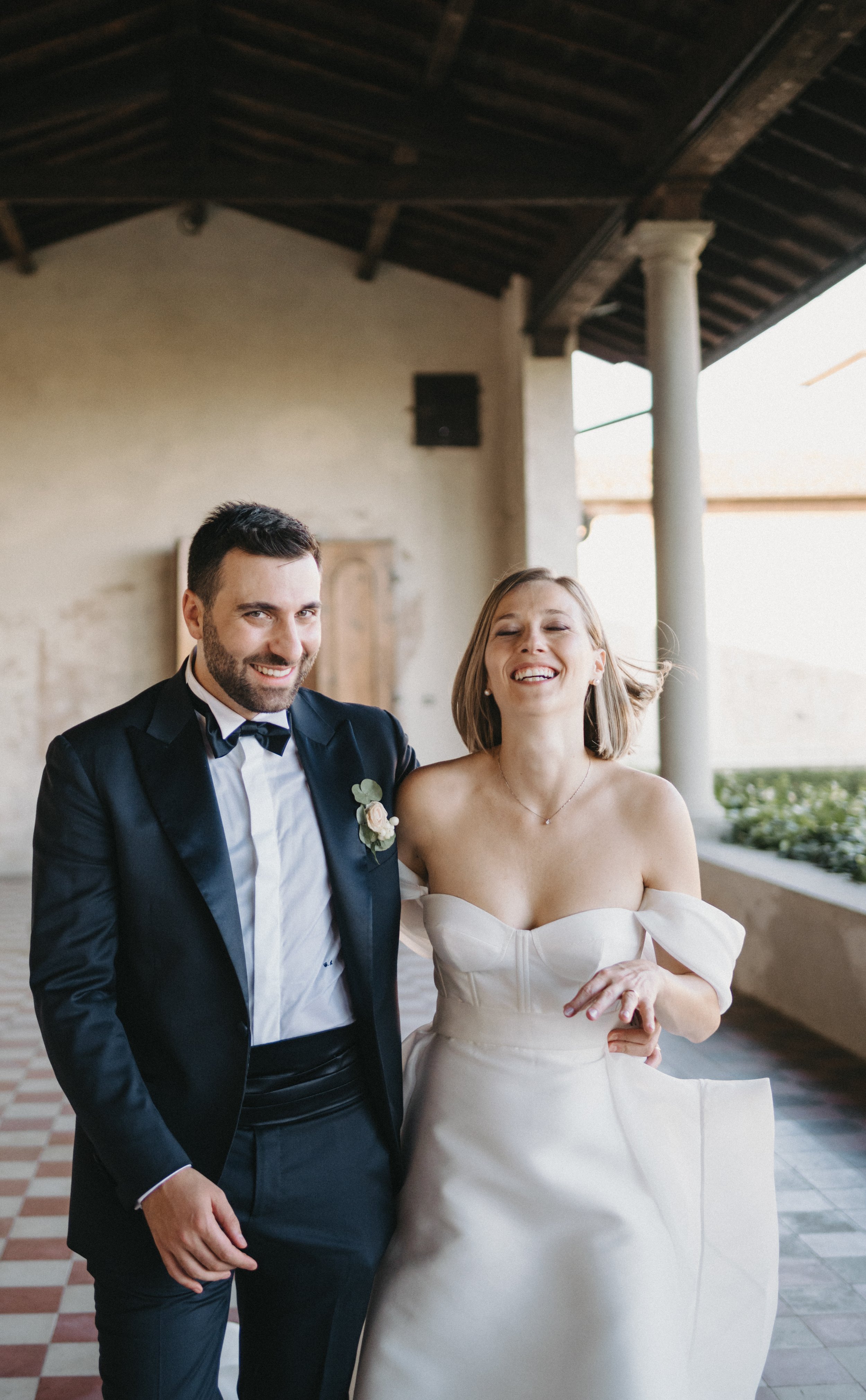 Una coppia sorridente in abiti da sposa, con lo sposo in completo nero e la sposa in abito bianco, in un ambiente coperto con colonne.