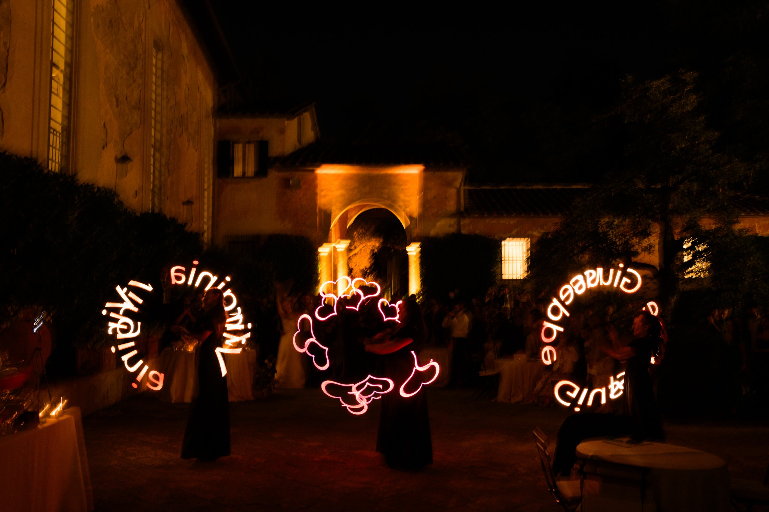 Spettacolo di luci con scritte luminose rosse in un cortile di sera, con sfondo di edifici storici e pubblico.