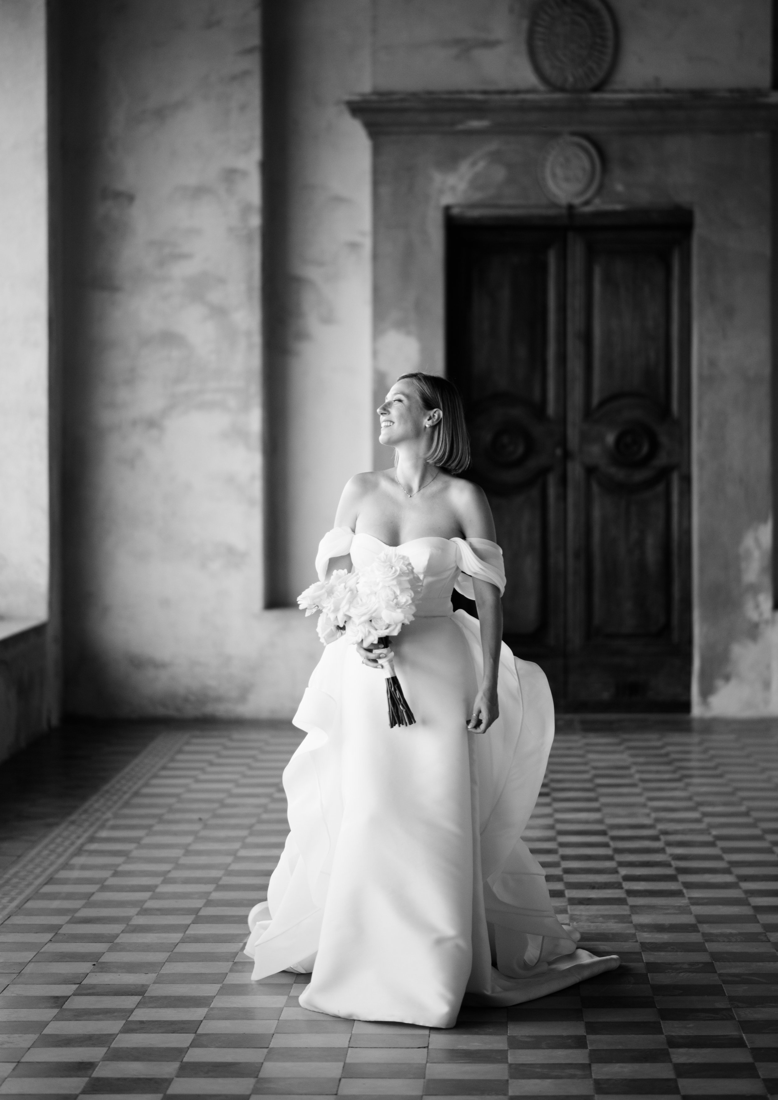 Sposa sorridente in abito bianco elegante con bouquet in mano, posizionata in un corridoio con pavimento a scacchi e porta antica sullo sfondo.