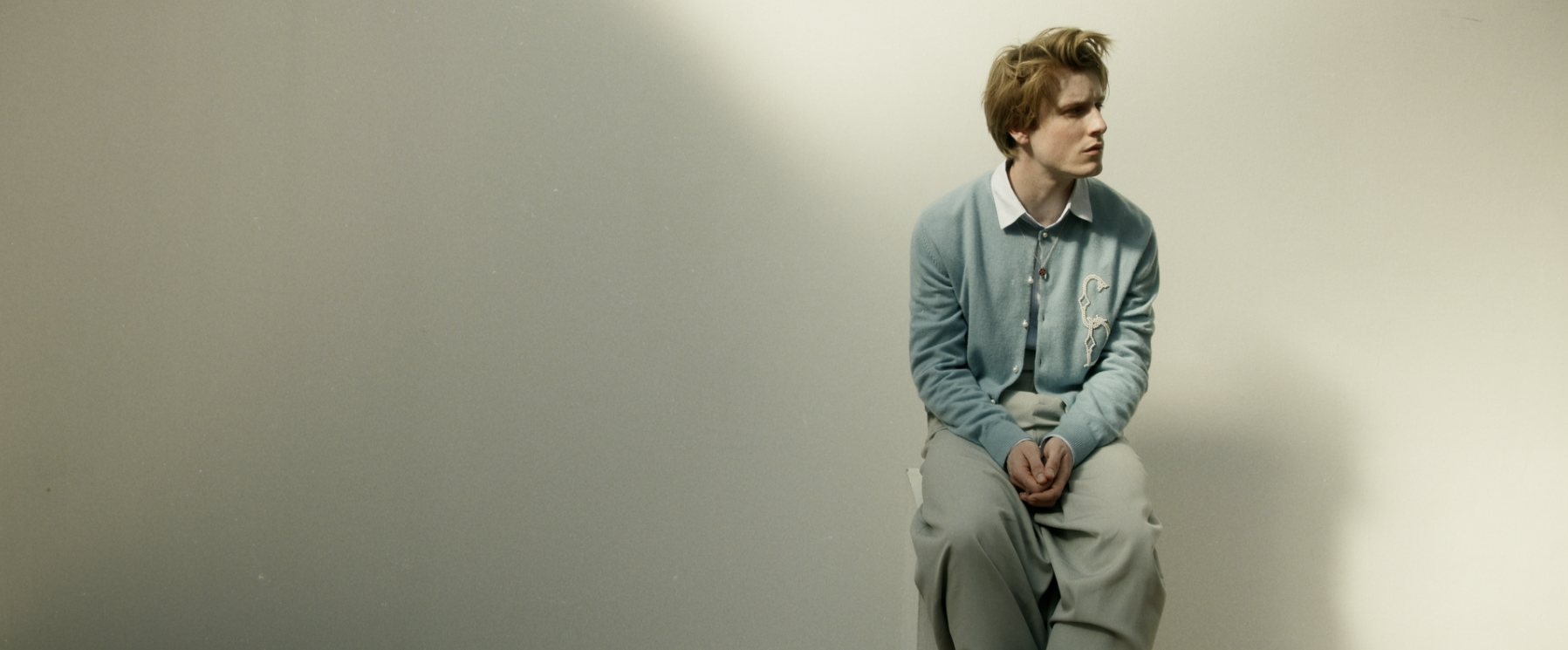Person sitting on a stool against a plain wall, wearing a light blue cardigan and light-colored pants.