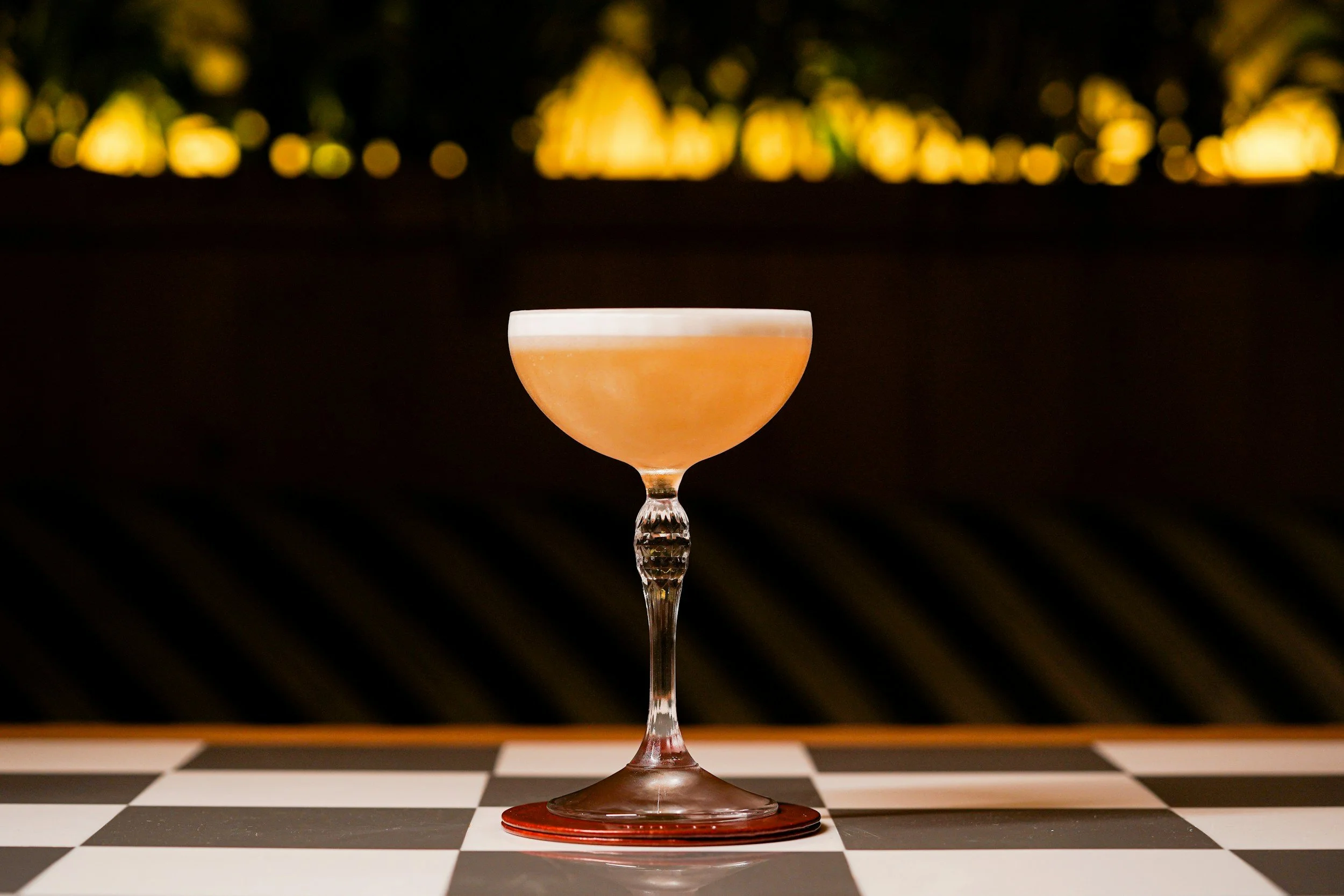 A cocktail drink in a coupe glass with a frothy white top, placed on a checkered black and white surface with blurred lights in the background.