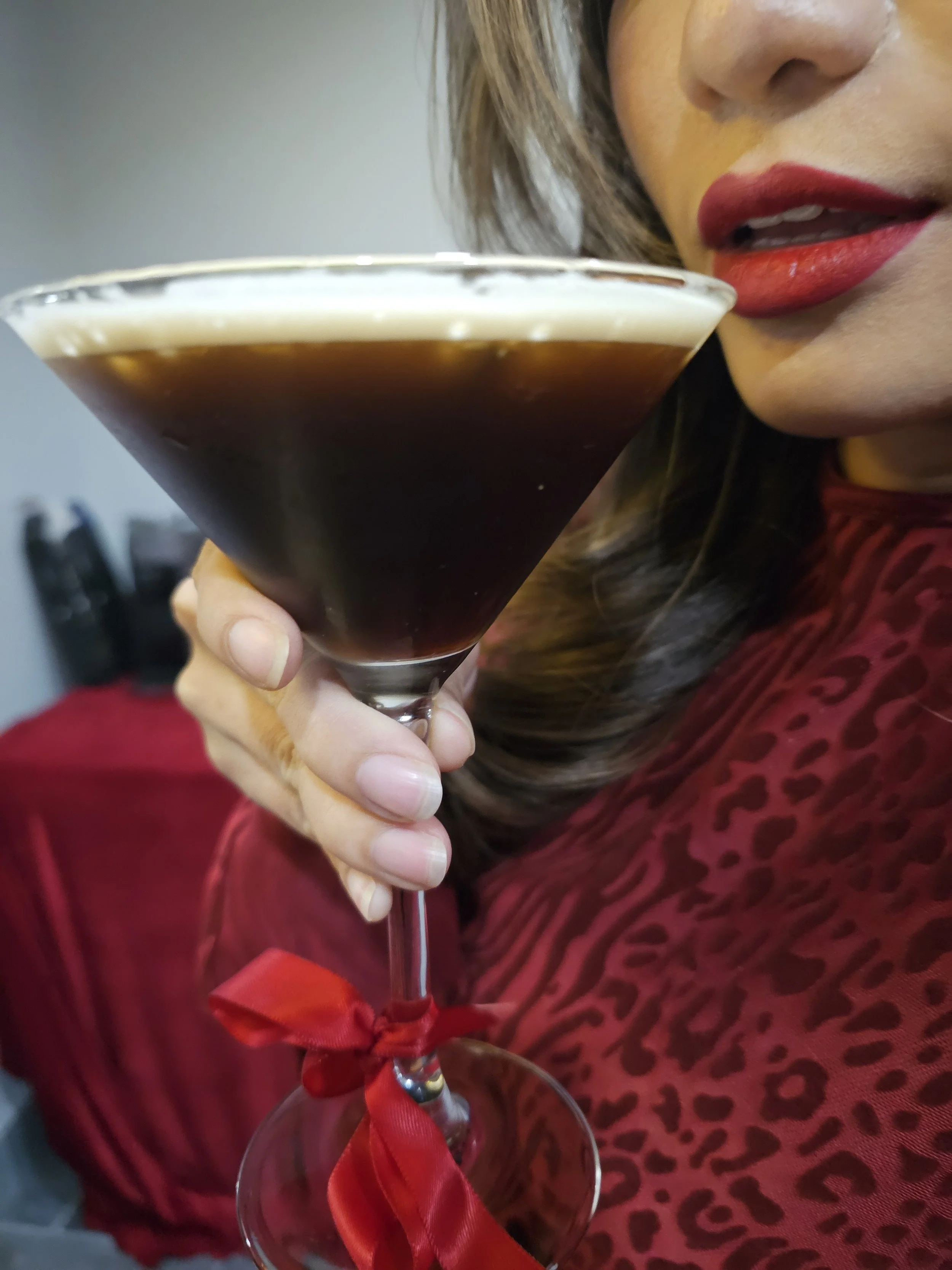 Close-up of a woman with red lipstick holding a cocktail glass with a dark beverage, tied with a red ribbon, and wearing a red leopard print outfit.