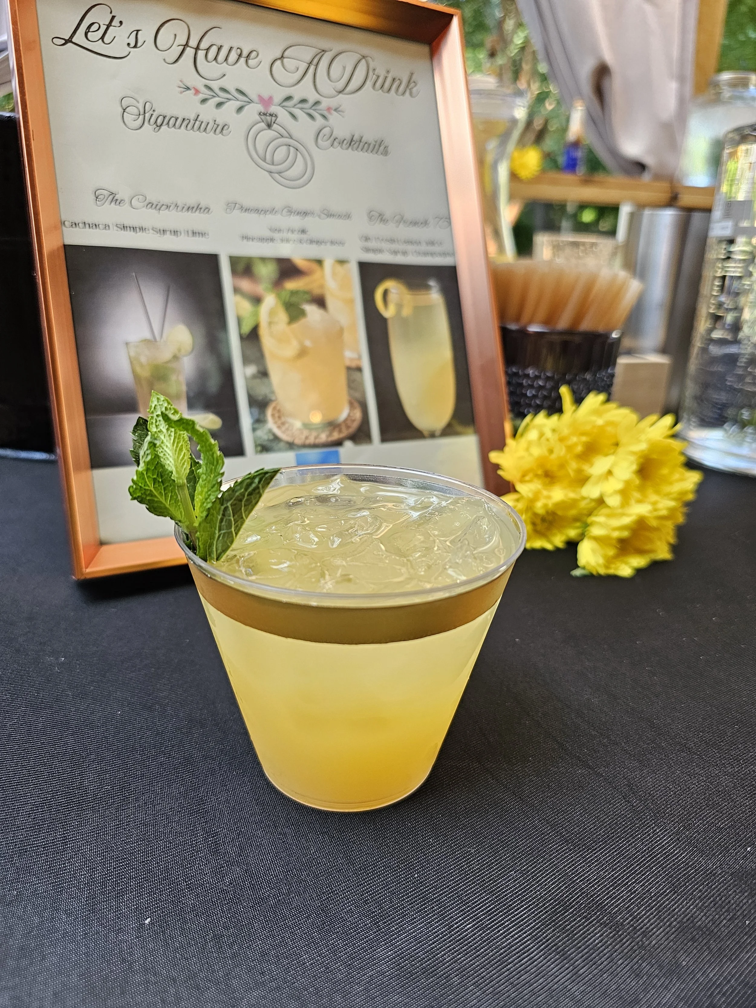 A yellow cocktail garnished with mint leaves on the rim, served in a clear glass with ice, placed on a black table with a yellow flower arrangement and a framed menu in the background.