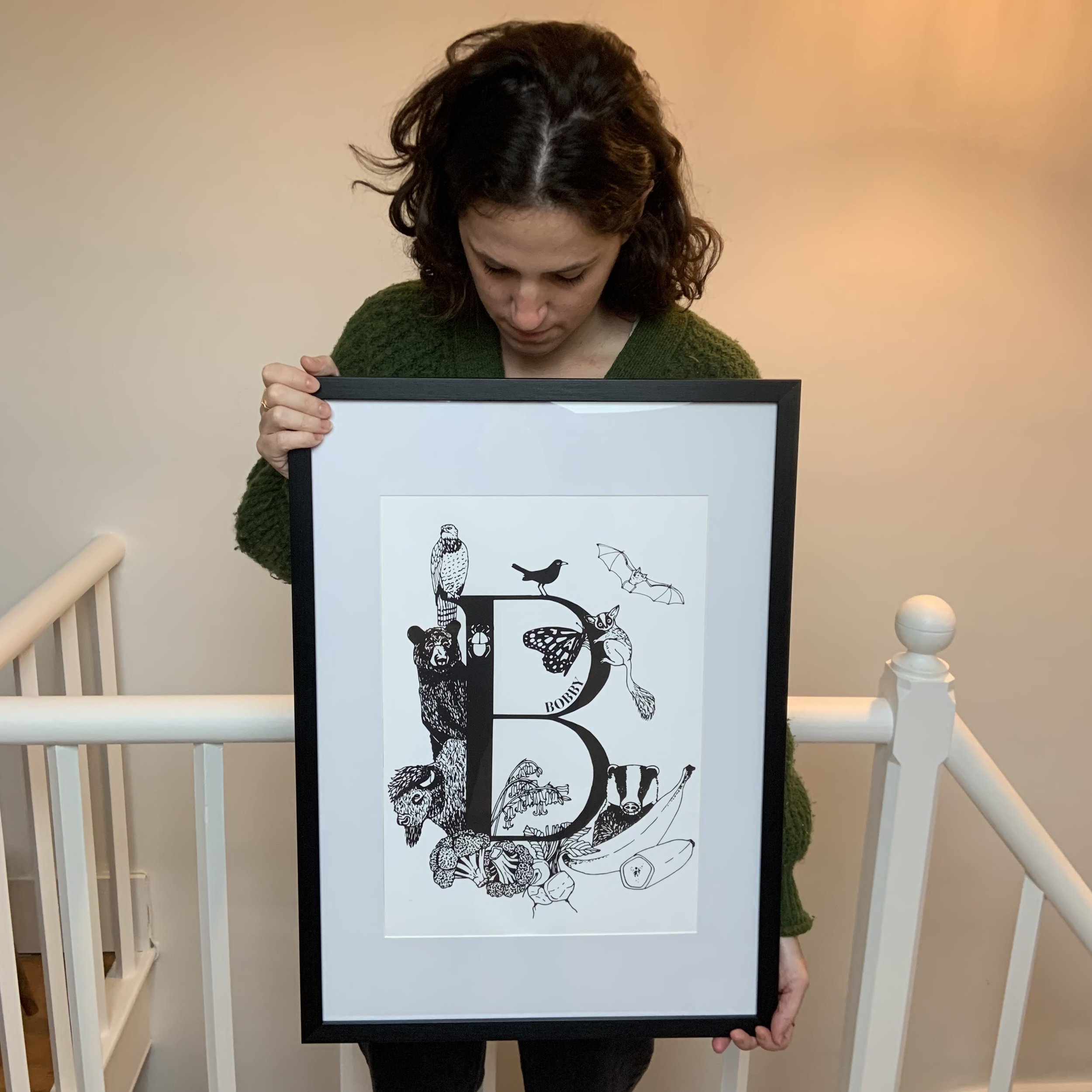 A person holding a framed black and white artwork featuring the letter 'B' surrounded by illustrations of various animals like birds, bats, and a bear.