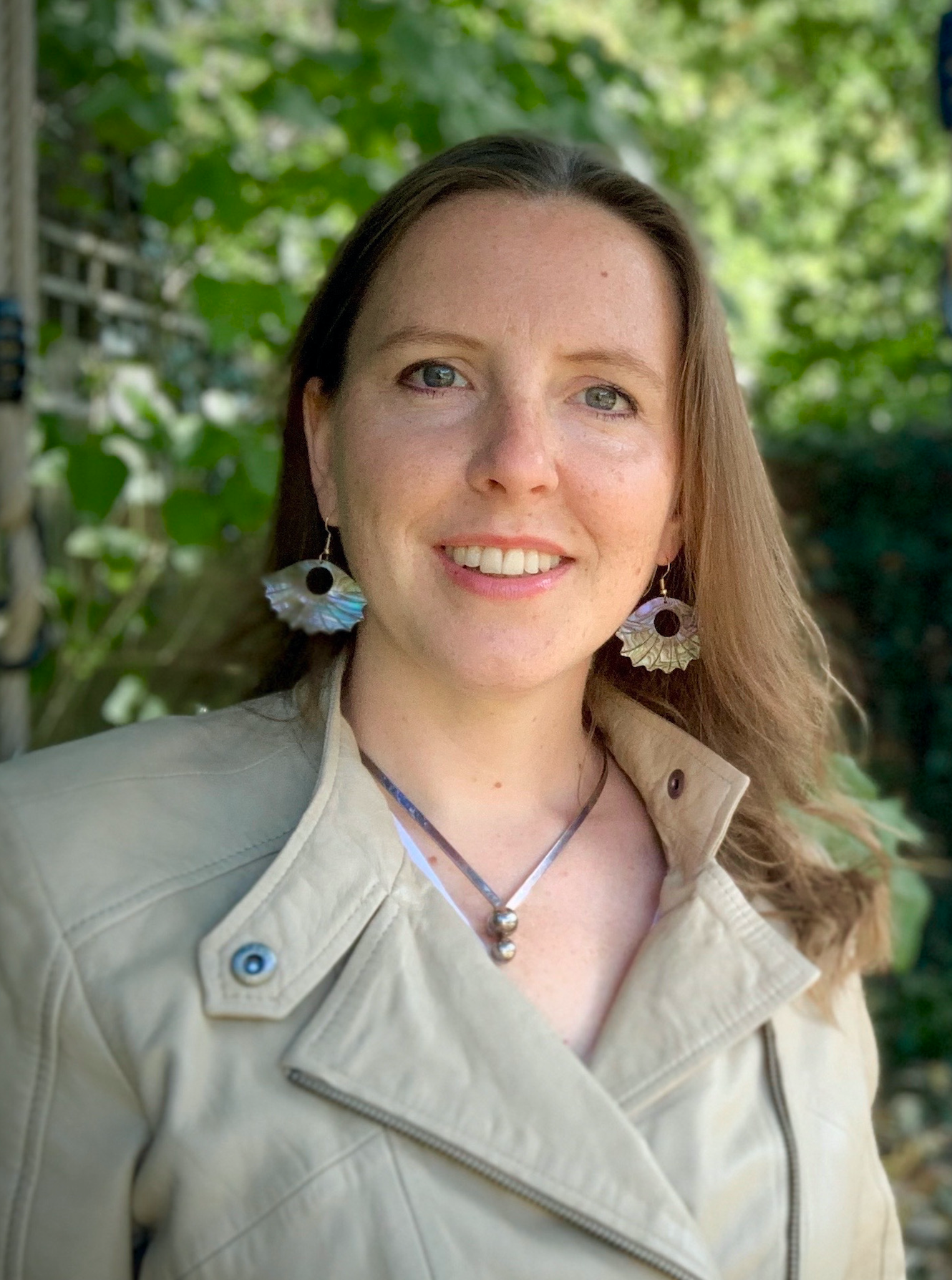 Wendy Lewis with brown hai, and blue eyes smiling outdoors with green trees in the background, wearing large earrings, a necklace with two silver beads, and a beige leather jacket.