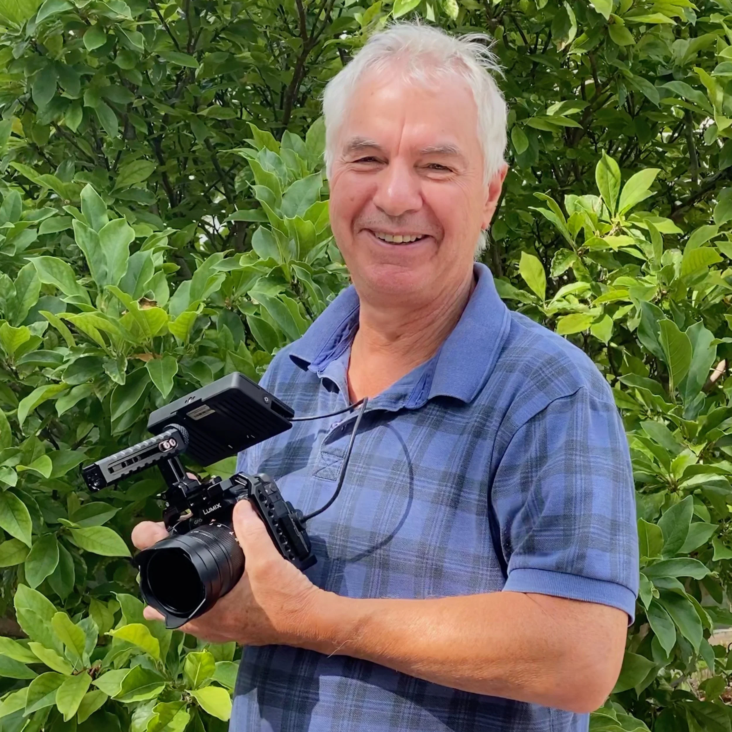 A smiling Rob Dunford with white hair holding a camera outdoors in front of green leafy plants.
