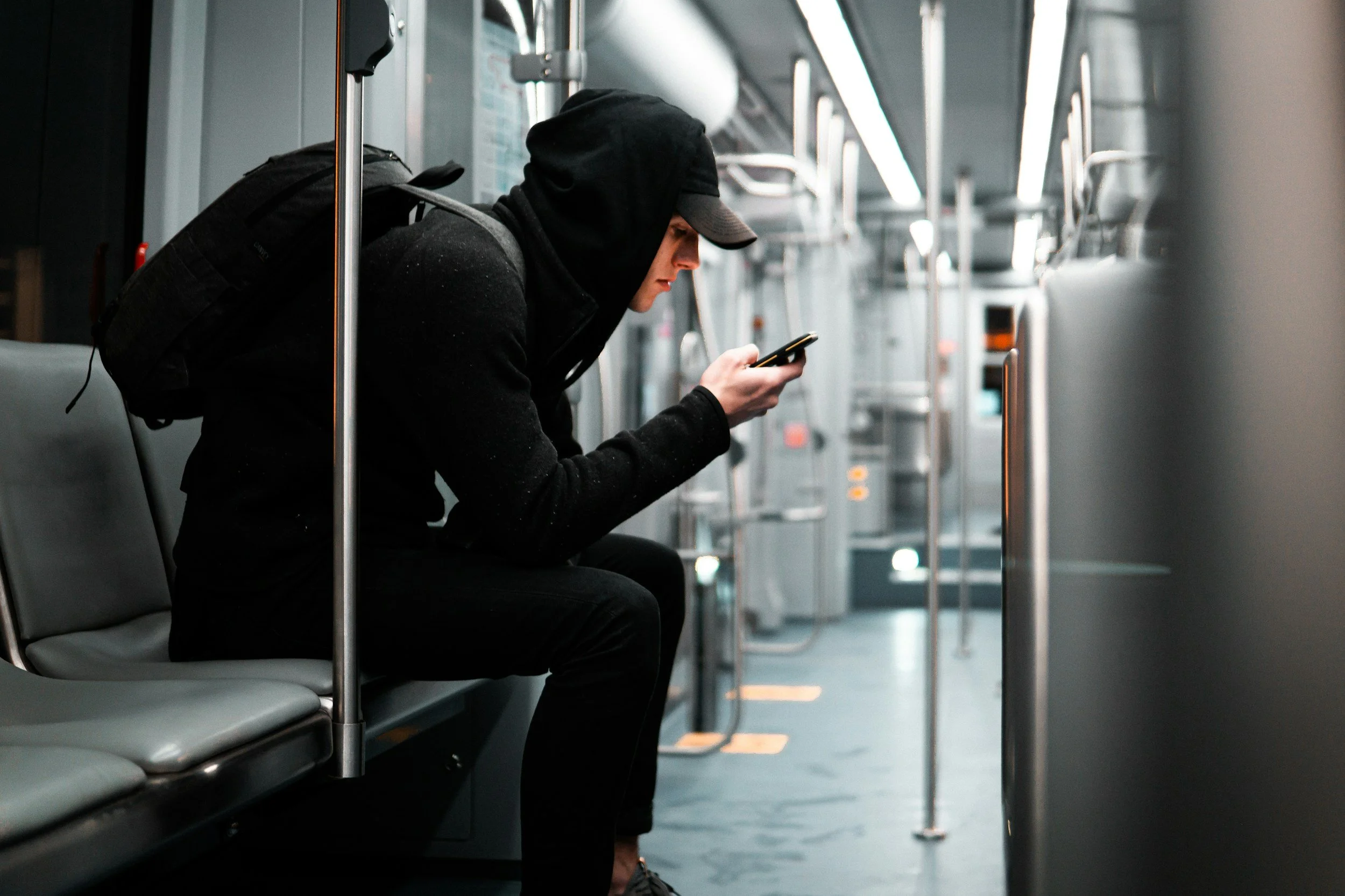 A man dressed in black, wearing a hoodie and cap, sitting alone on a bench inside a mostly empty subway train, looking at his phone.