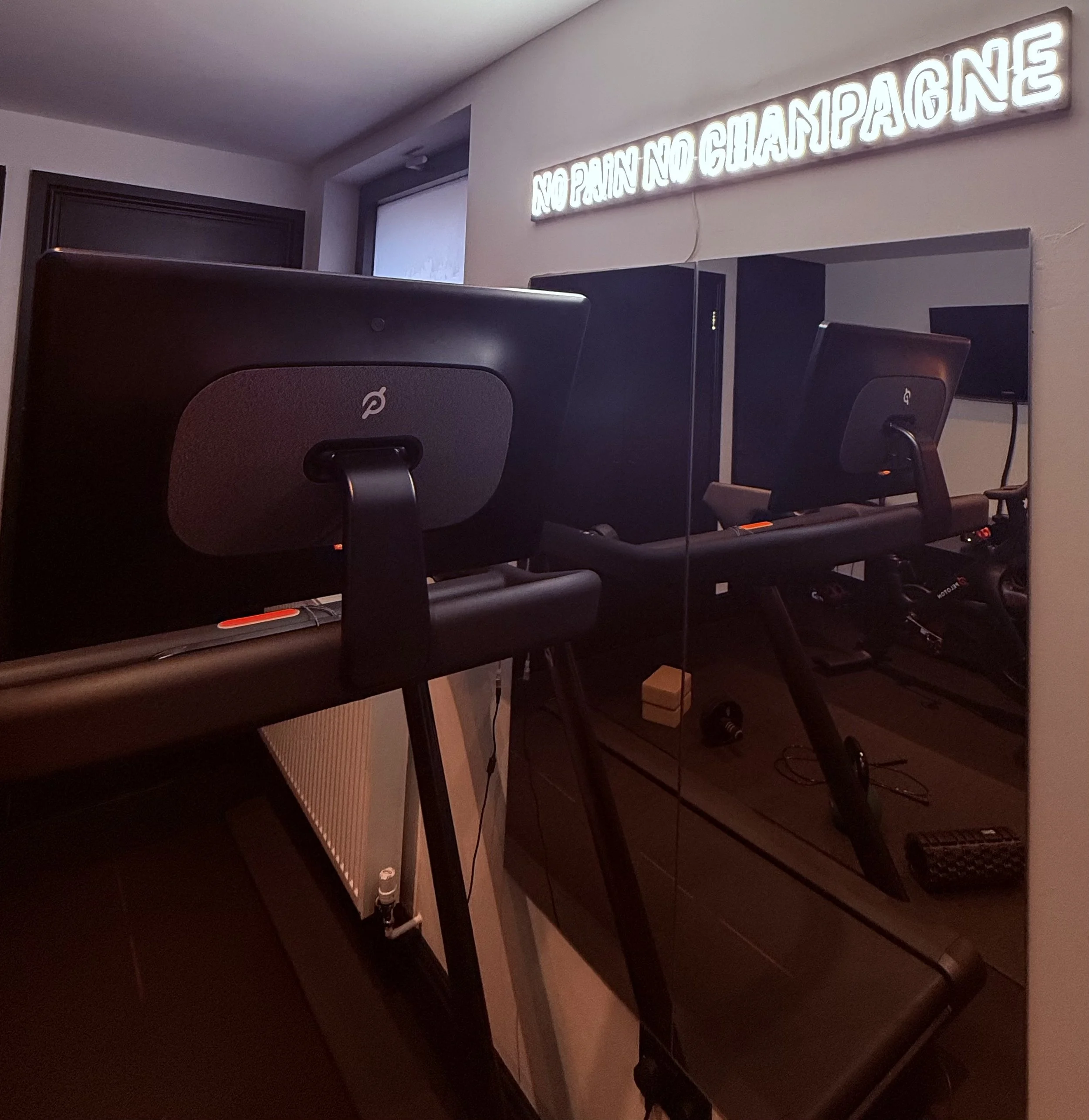 Dark-colored treadmill facing a mirror, with a neon sign on the wall that reads 'NO PRANK NO CHAMPAGNE' in a dimly lit room.
