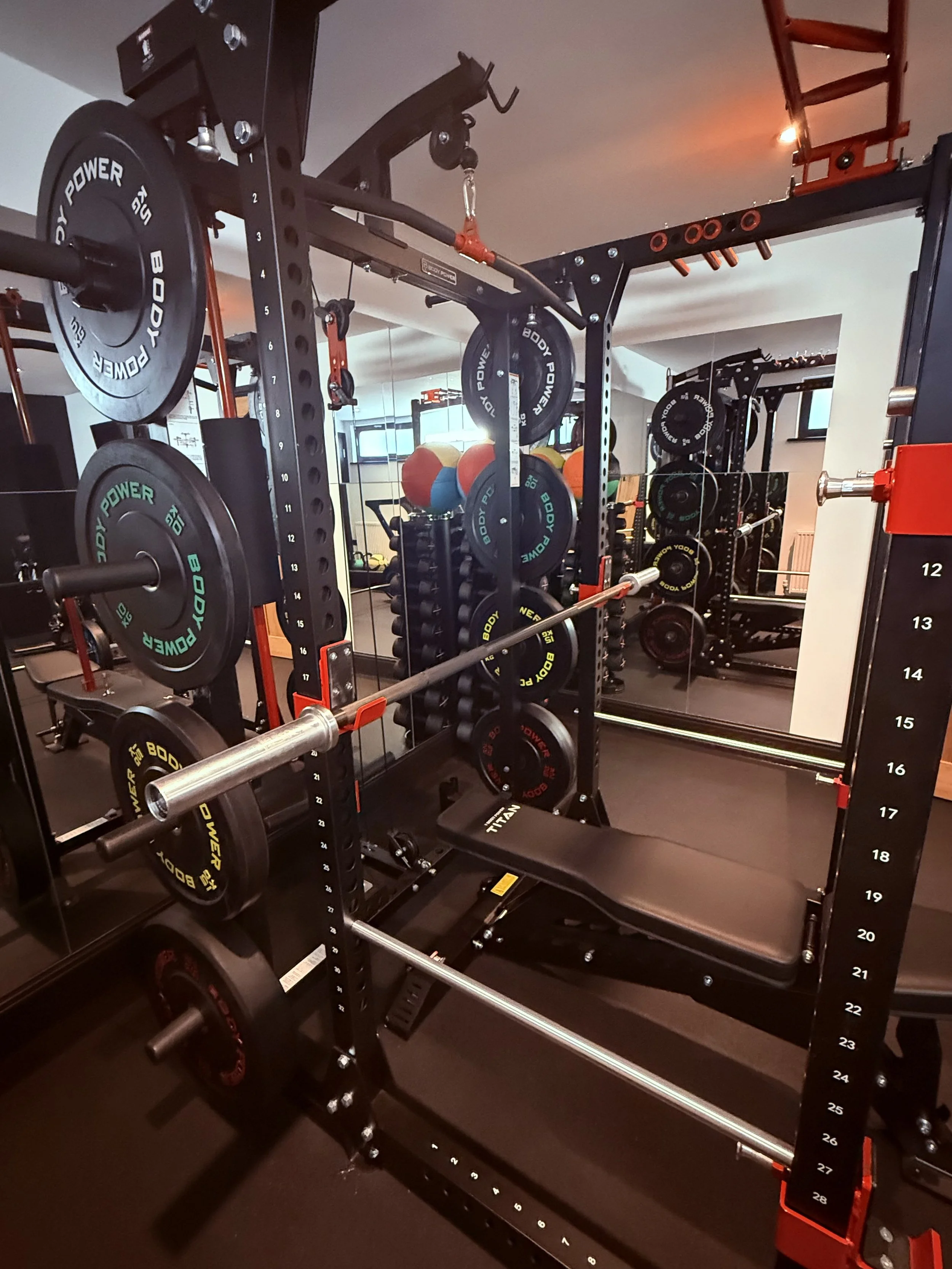 A gym weightlifting station with a black power rack, adjustable barbell, weight plates, and mirror. The weights include black, green, and yellow plates labeled 'BODY POWER 5KG.'