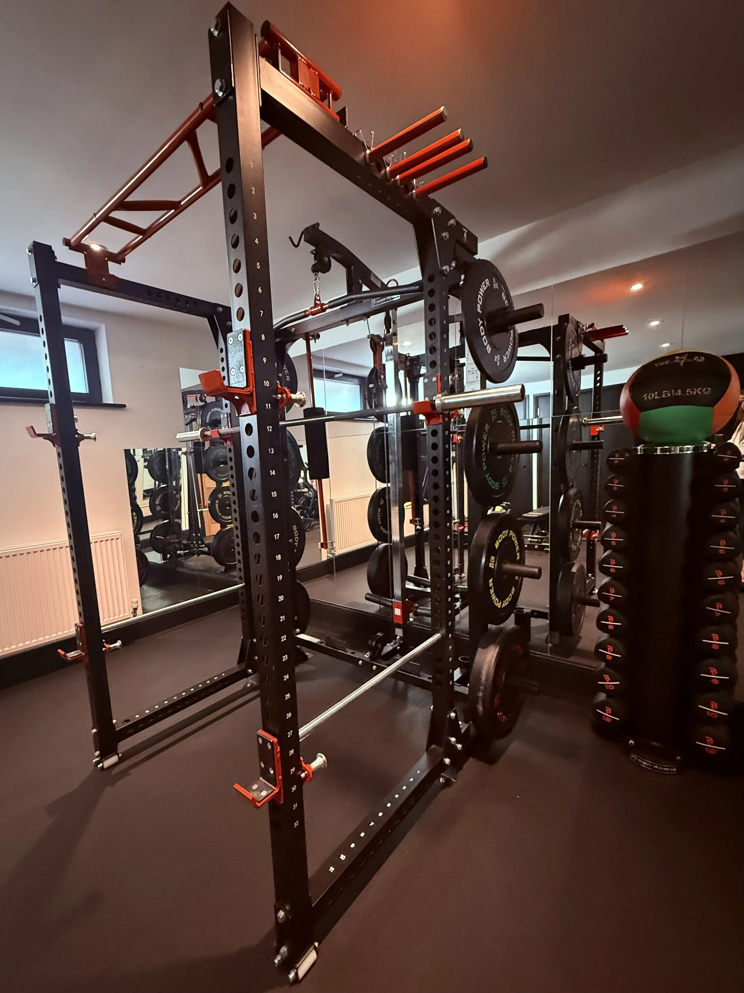 Indoor gym with various weightlifting equipment, including a squat rack, barbells, weight plates, and a stack of dumbbells. The room has mirrors and a window.