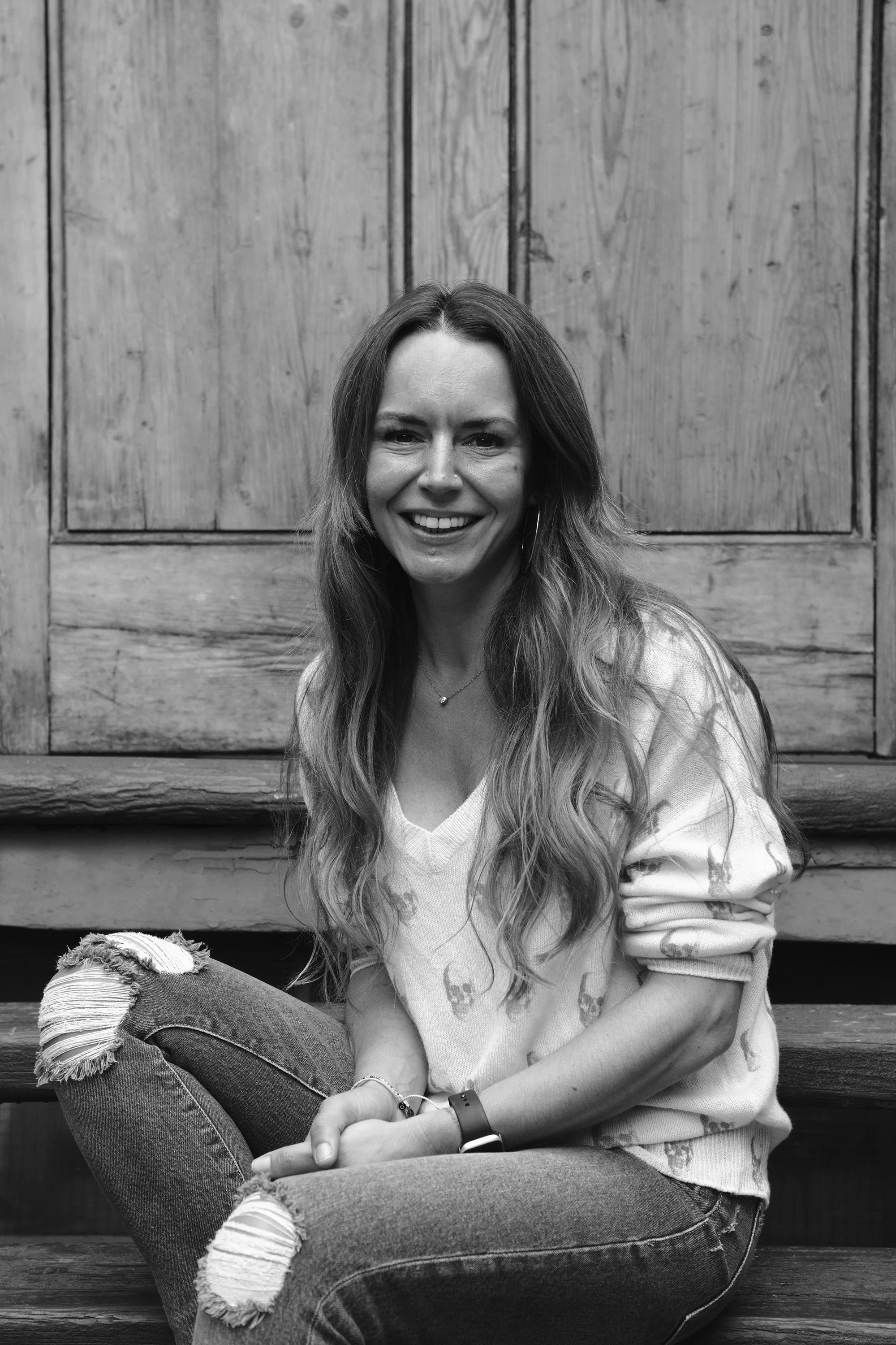 A woman with long wavy hair sitting on wooden stairs, smiling at the camera, wearing a sweater with skull prints, ripped jeans, and accessories.