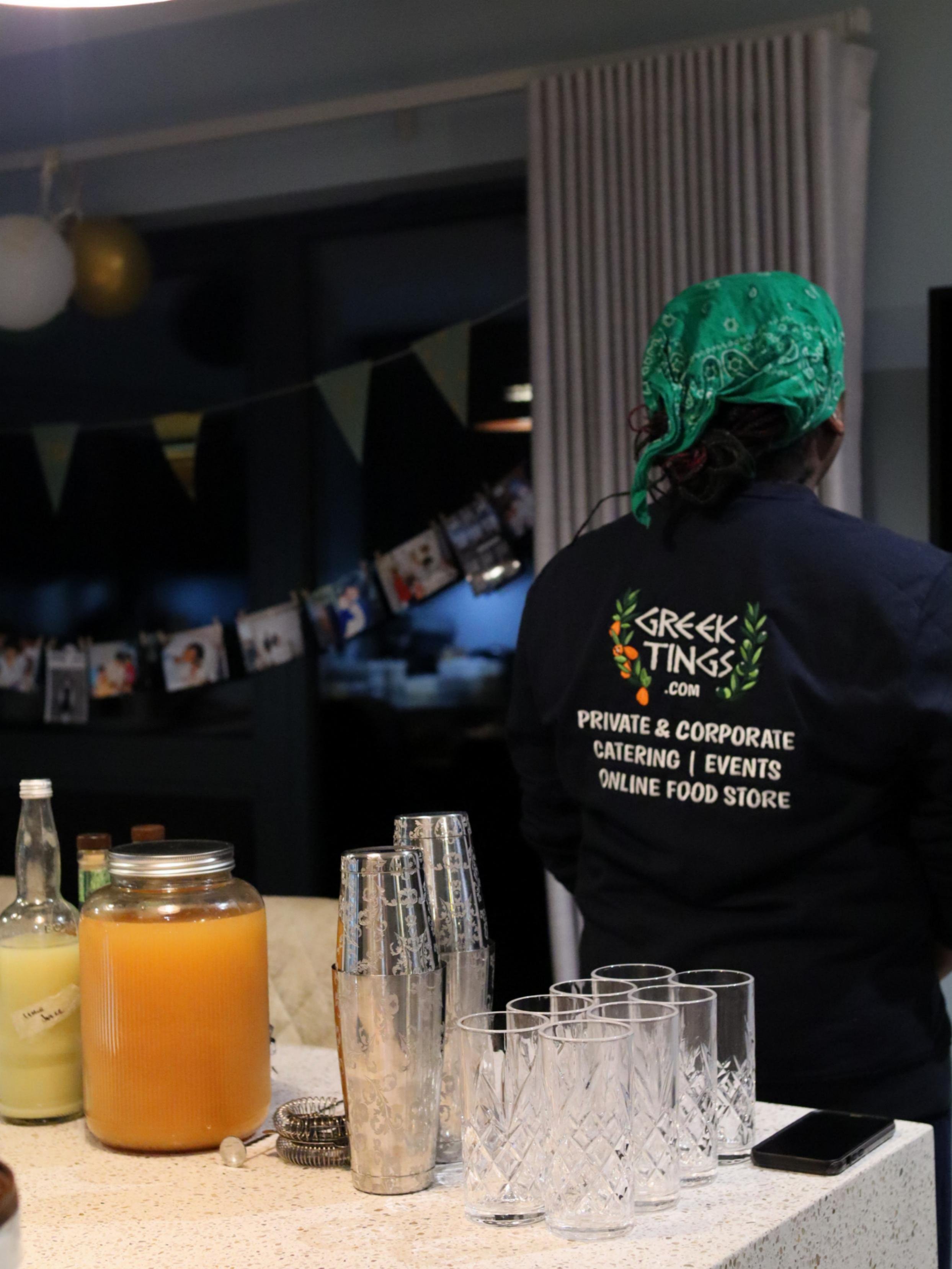 Greek Tings team member preparing drinks beside cocktail shakers and glassware at an event.
