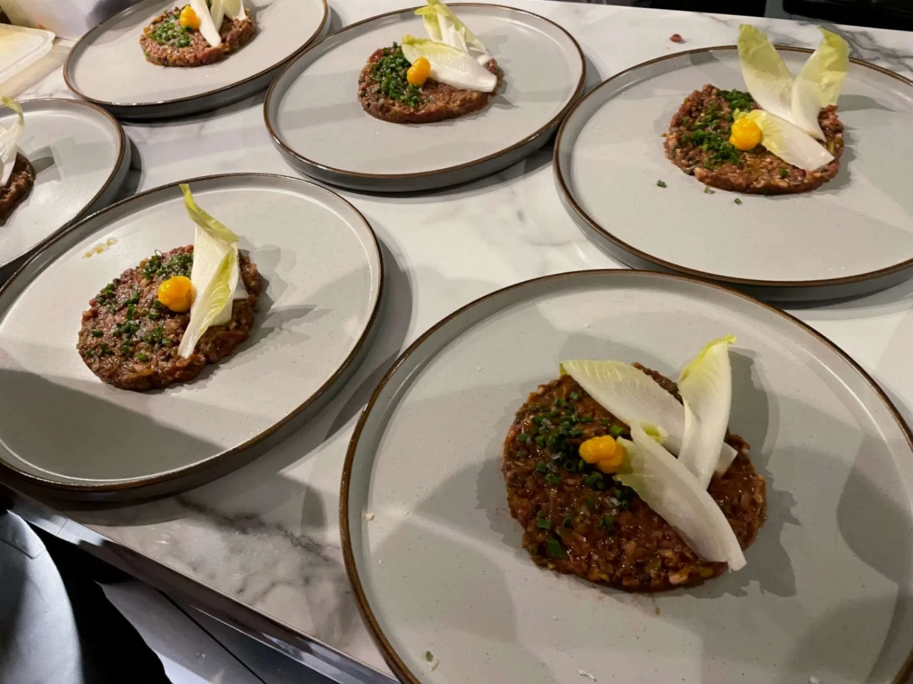 Plated beef tartare starters topped with chives, endive leaves and a yellow condiment.
