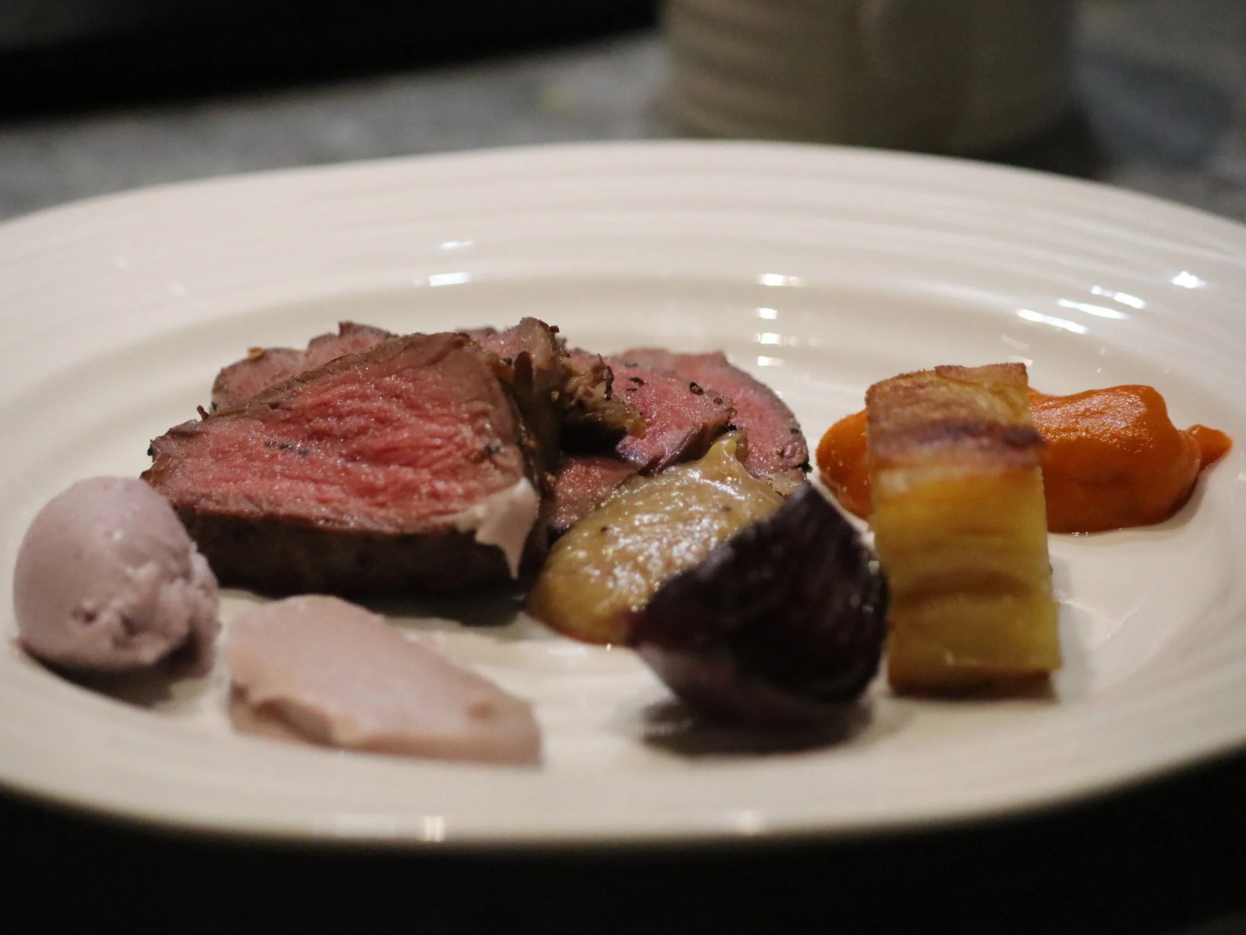 Fine dining roast meat course with orange purée, dark vegetables and layered potato terrine.
