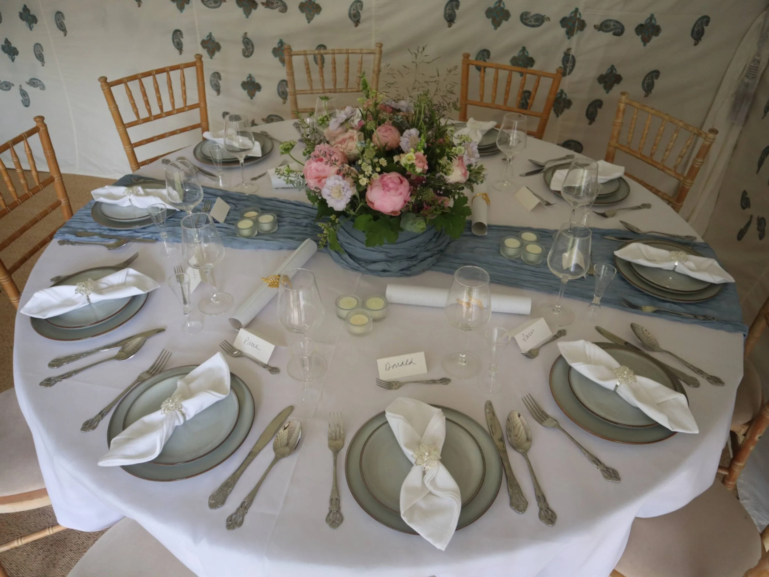  Round private dining table with blue runner, floral centrepiece, candles and formal place settings.
