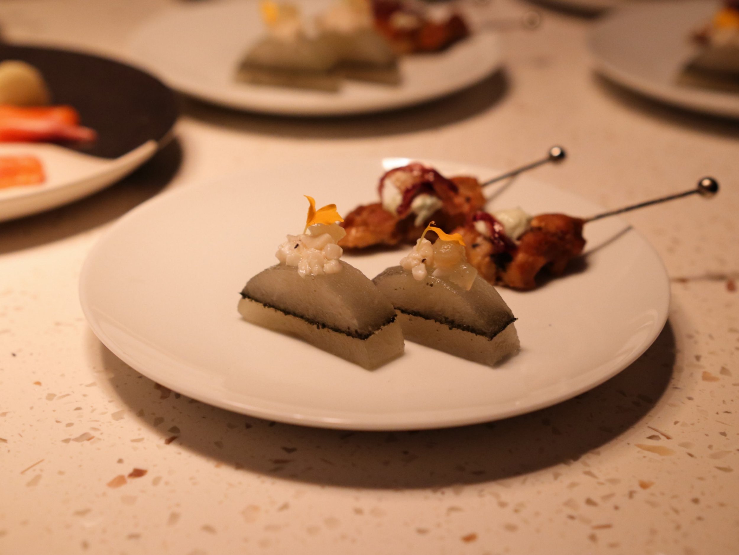 Fine dining tasting plate with delicate layered bites and skewered savoury pieces.