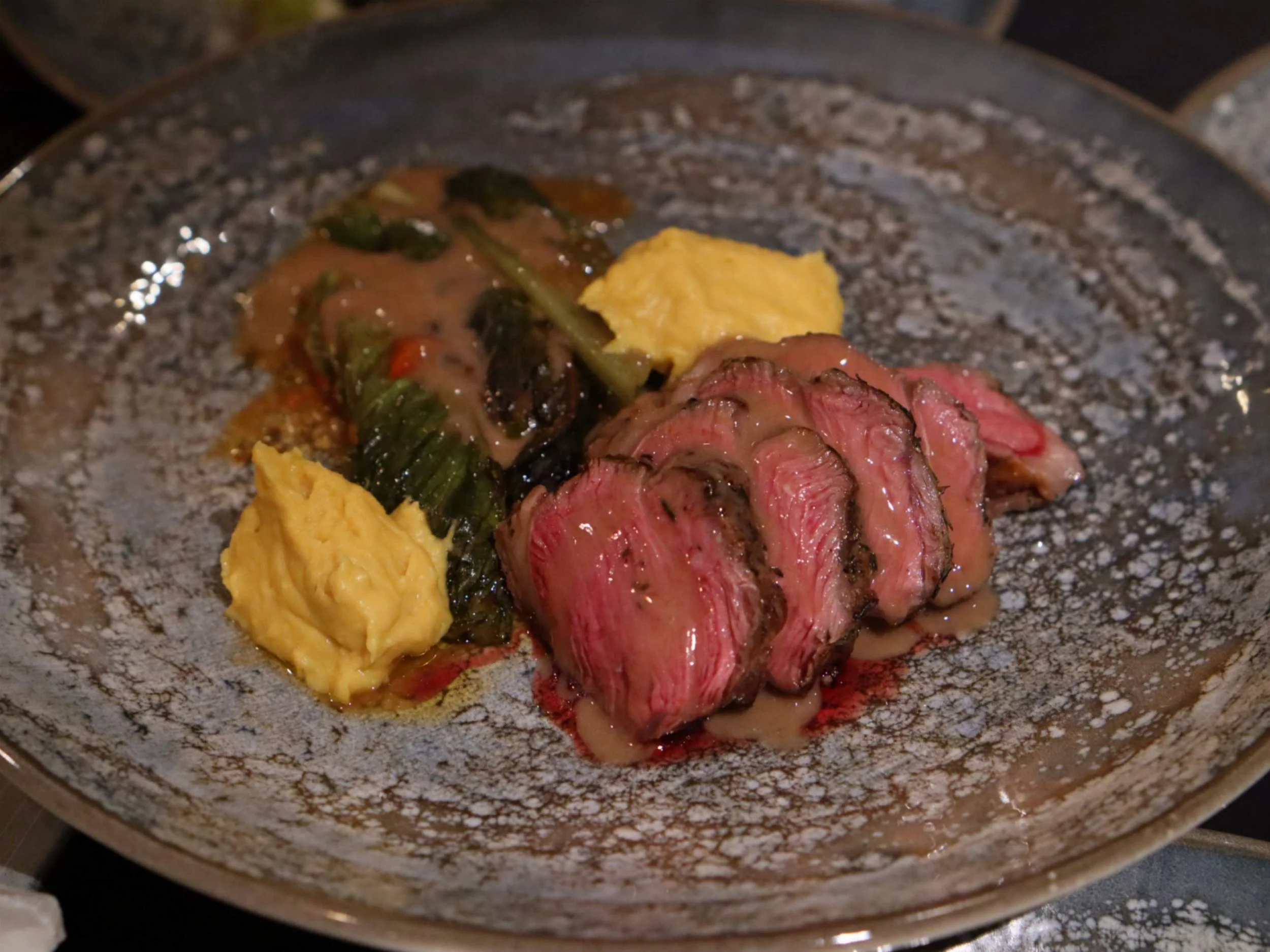 Pink roast meat with greens, yellow purée and rich sauce on a fine dining plate.
