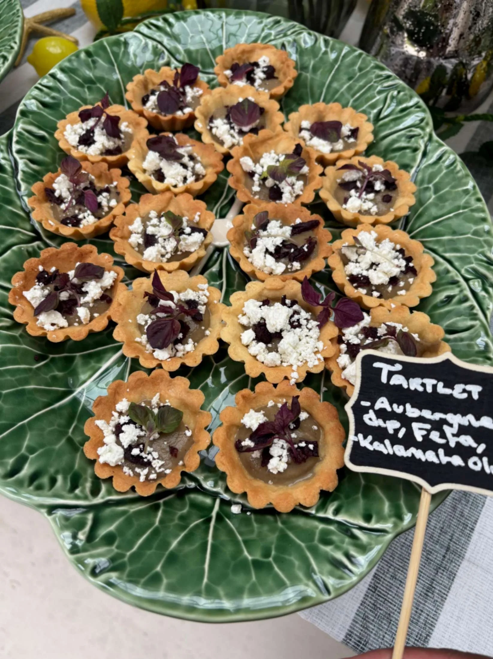 Mini aubergine and feta tartlets with Kalamata olive and micro herbs on a green serving platter.
