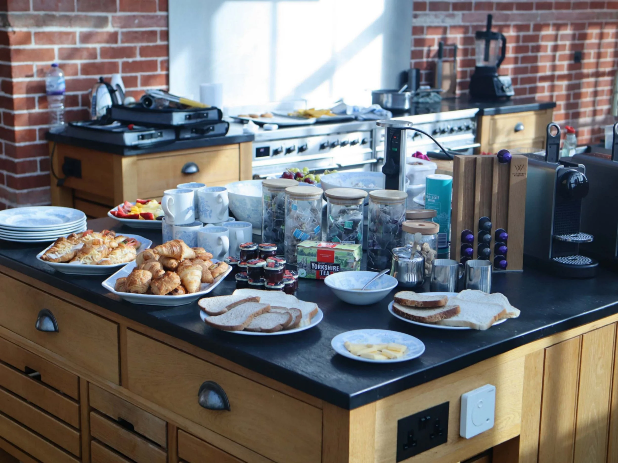 Private breakfast buffet with croissants, toast, preserves, tea and coffee on a kitchen island.
