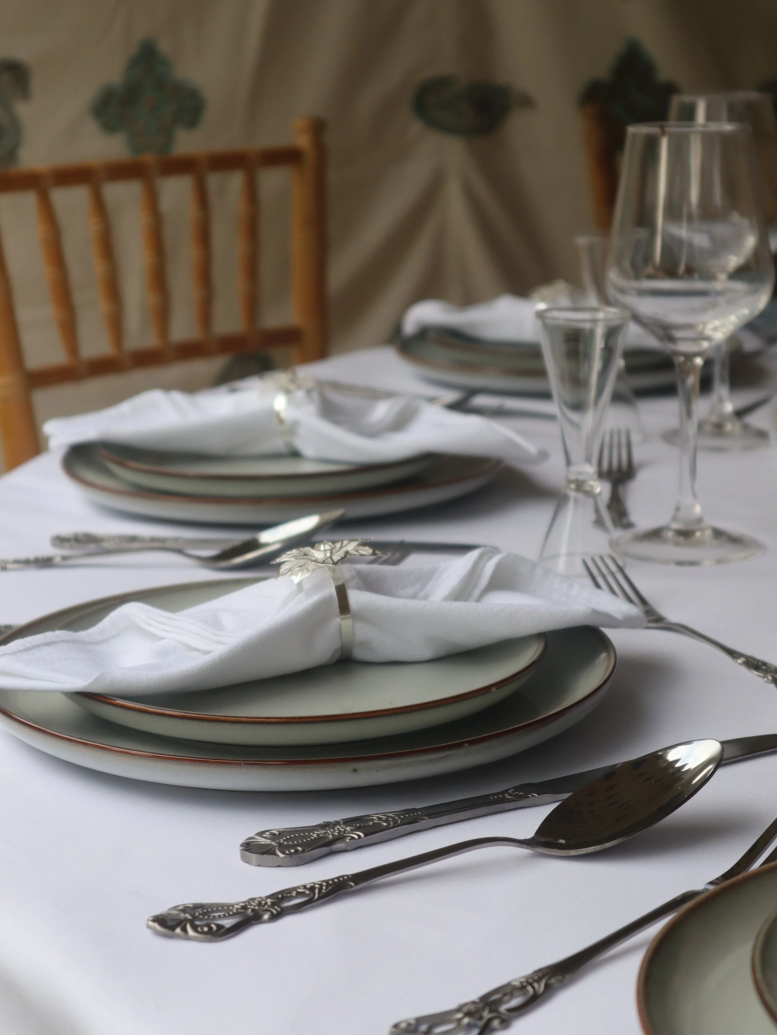 Table setting, large charger plate with smaller starter plate on top,  fine dining, white napkin with silver floral napkin ring, silver ornate cutlery around plates, one large wine glass and one small shot glass next to it around every plate
