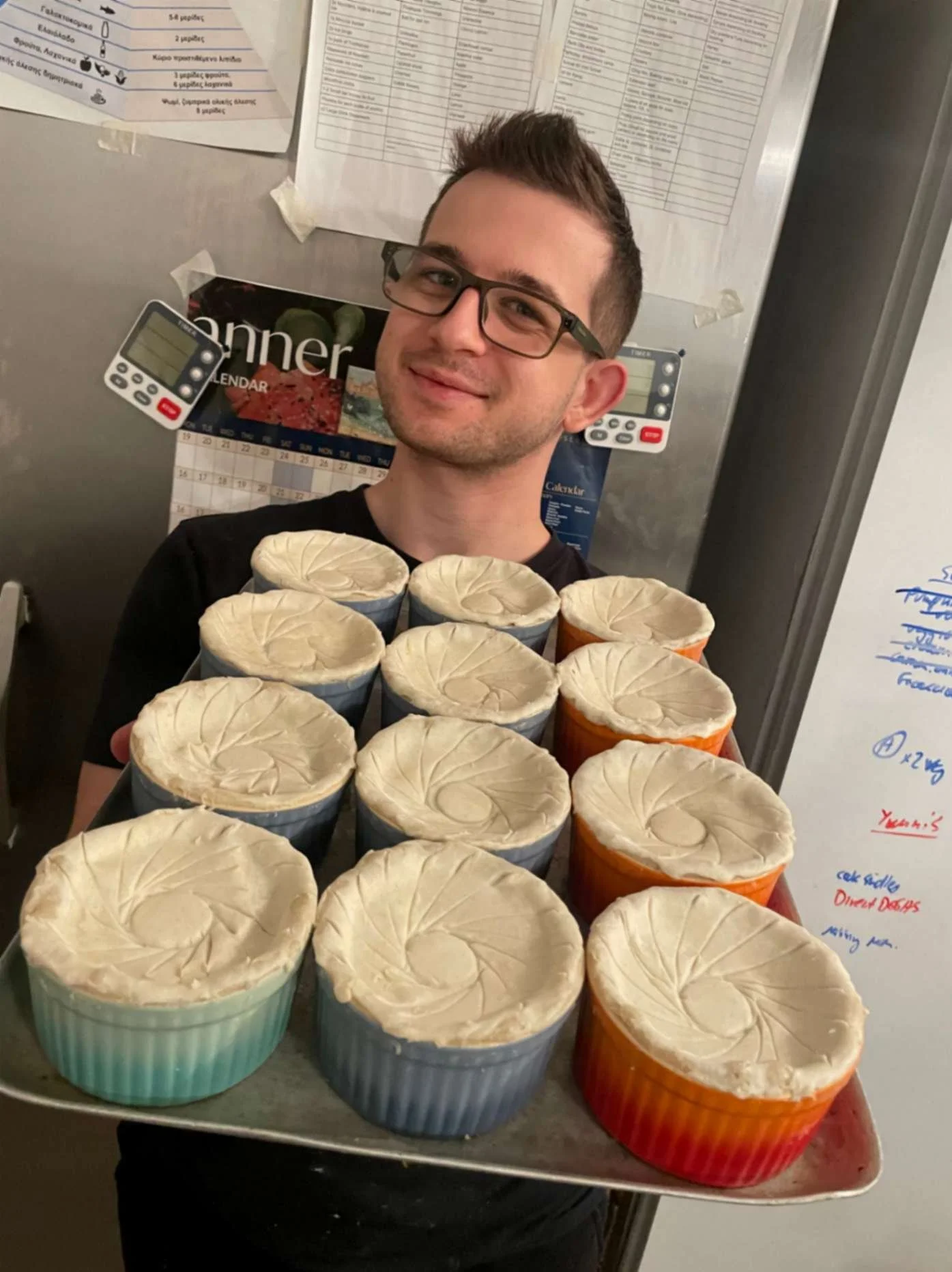 Greek Tings chef holding a tray of handmade pastry-topped pies in colourful ramekins.
