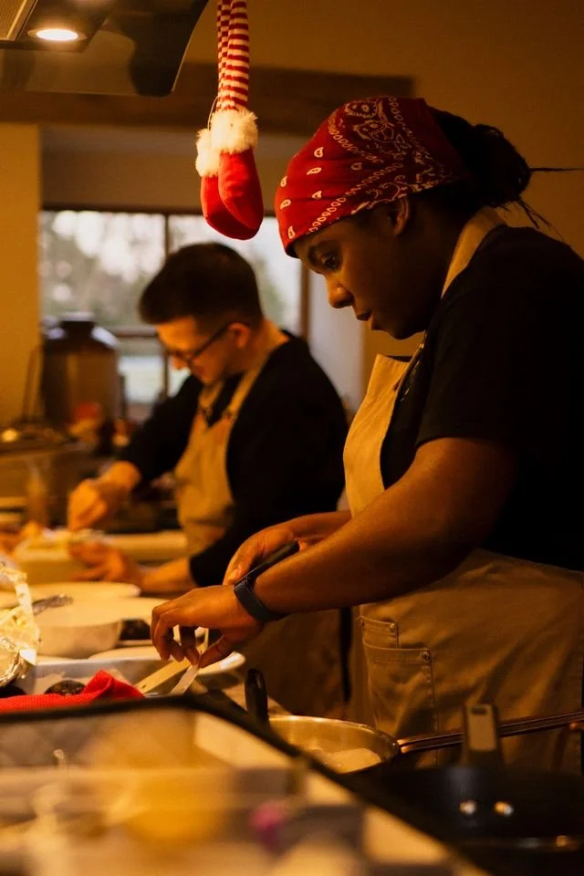 Two chefs, one greek man, one black woman wearing a red bandana, both plating food, sticky toffee pudding, vanilla ice cream
