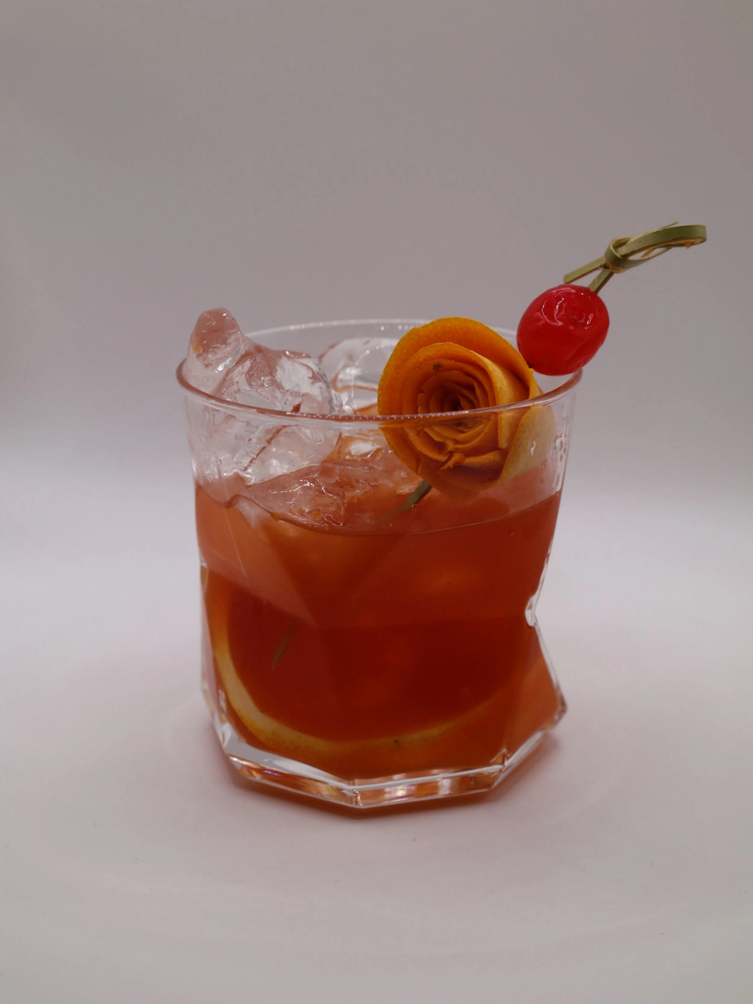 Amber cocktail over ice with a sculpted orange peel and cherry garnish.
