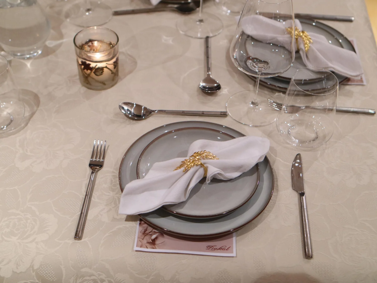 Formal place setting with grey plates, white napkin, gold wheat ring and glassware at a Greek Tings event.