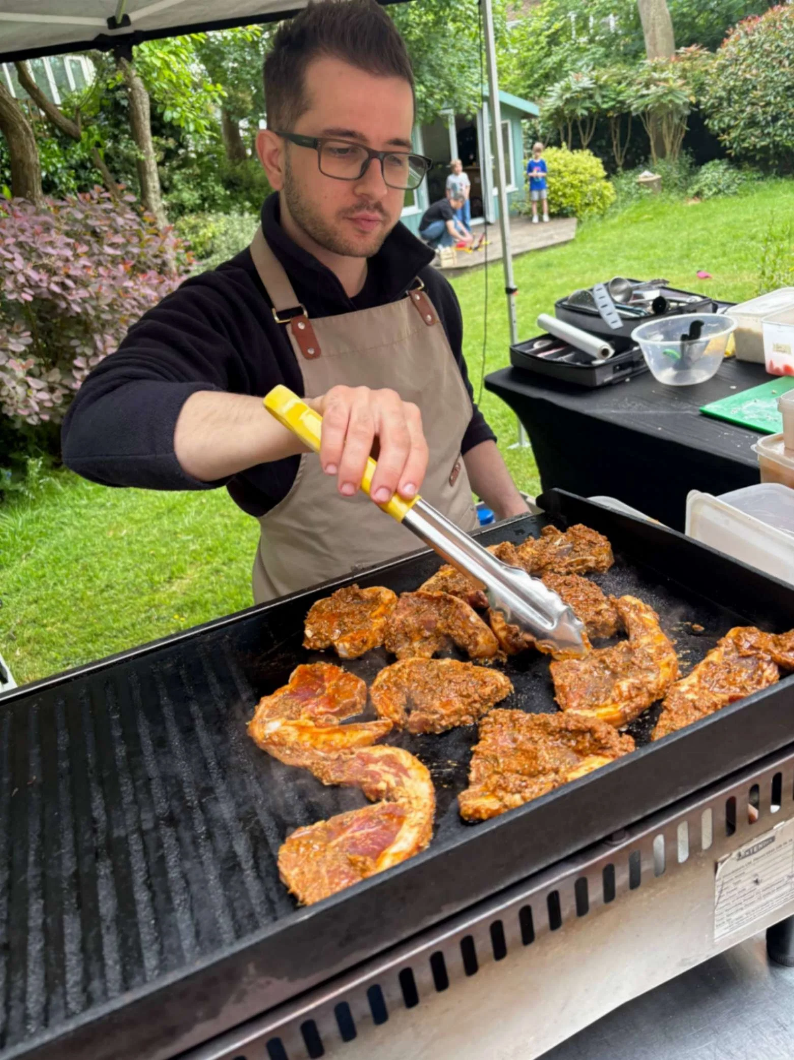 Greek Tings chef grilling marinated meat on a flat-top barbecue at a garden event.