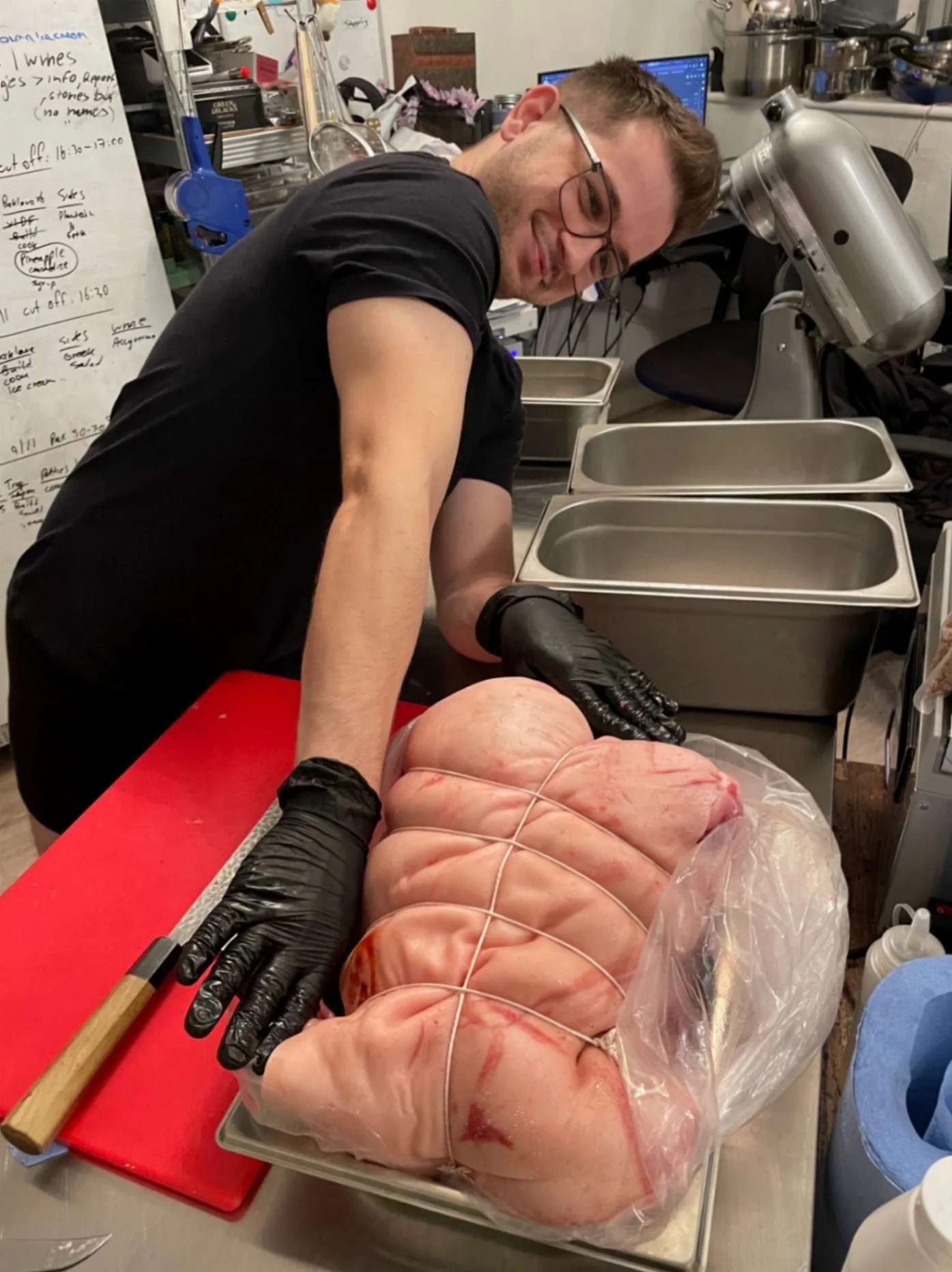 Greek Tings chef preparing a large tied pork roast on a red chopping board.
