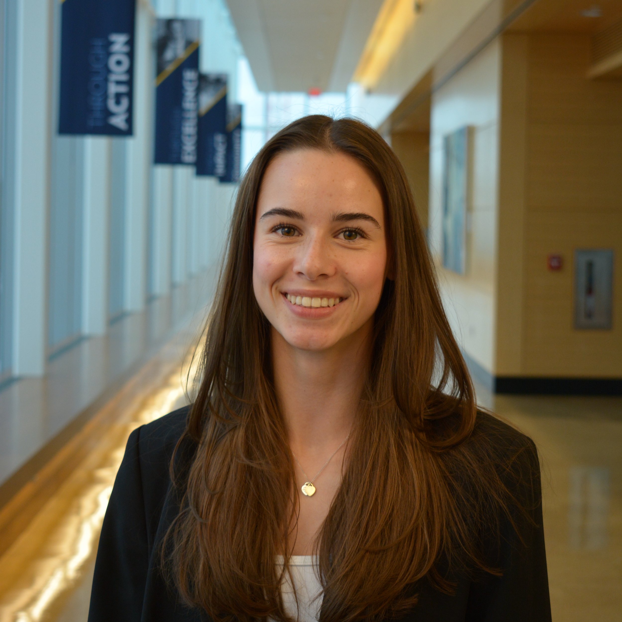 A young woman with long brown hair, smiling, wearing a black blazer and a necklace, standing in a modern building with banners hanging from the ceiling.