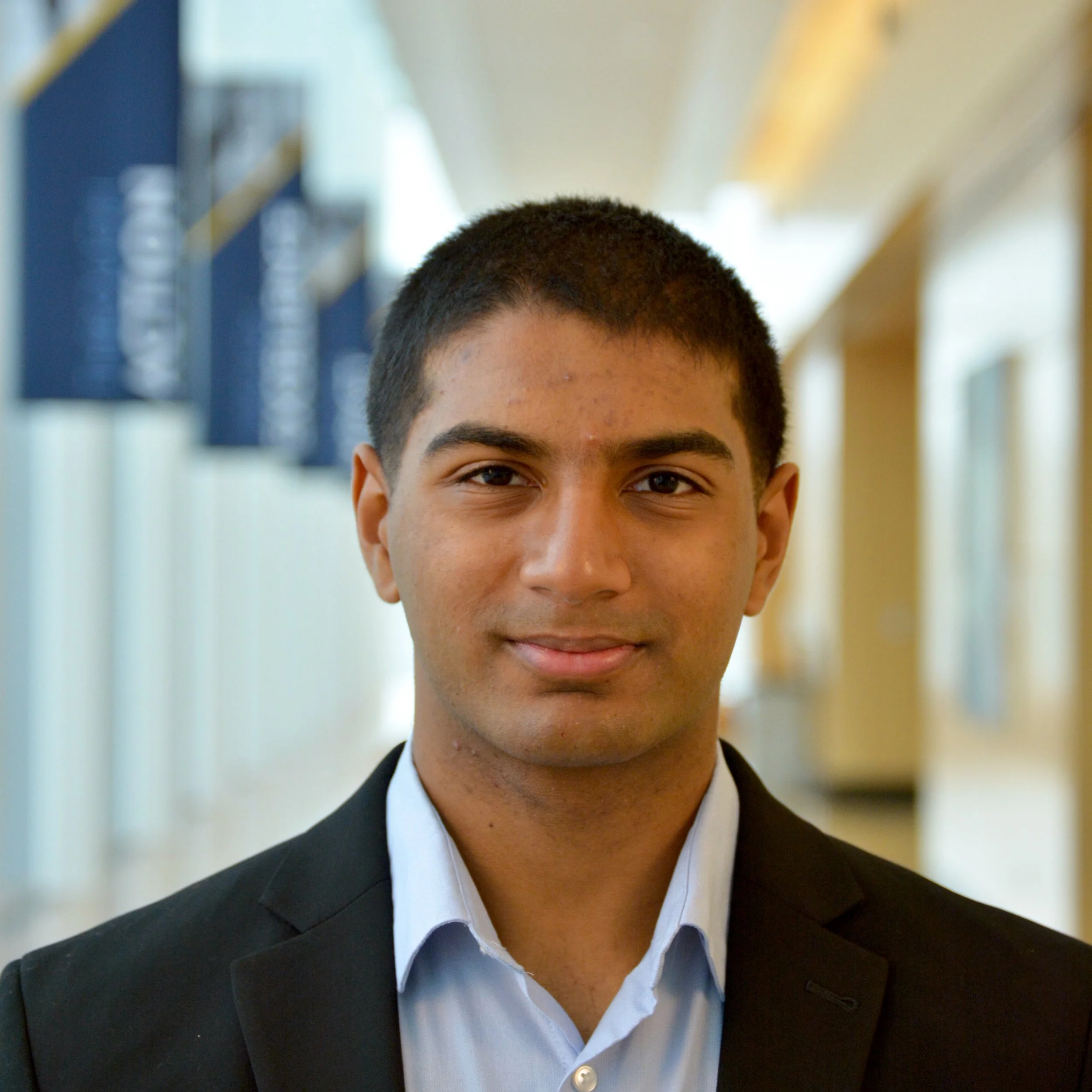Portrait of a young man with short black hair, wearing a suit and dress shirt, standing in a hallway with blurred background of windows and signs.