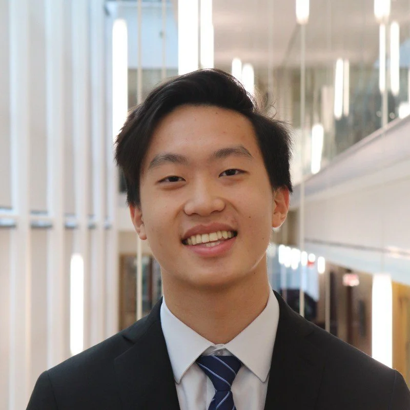 A young man in a black suit and striped tie smiling in a modern indoor setting with glass and lighting fixtures.