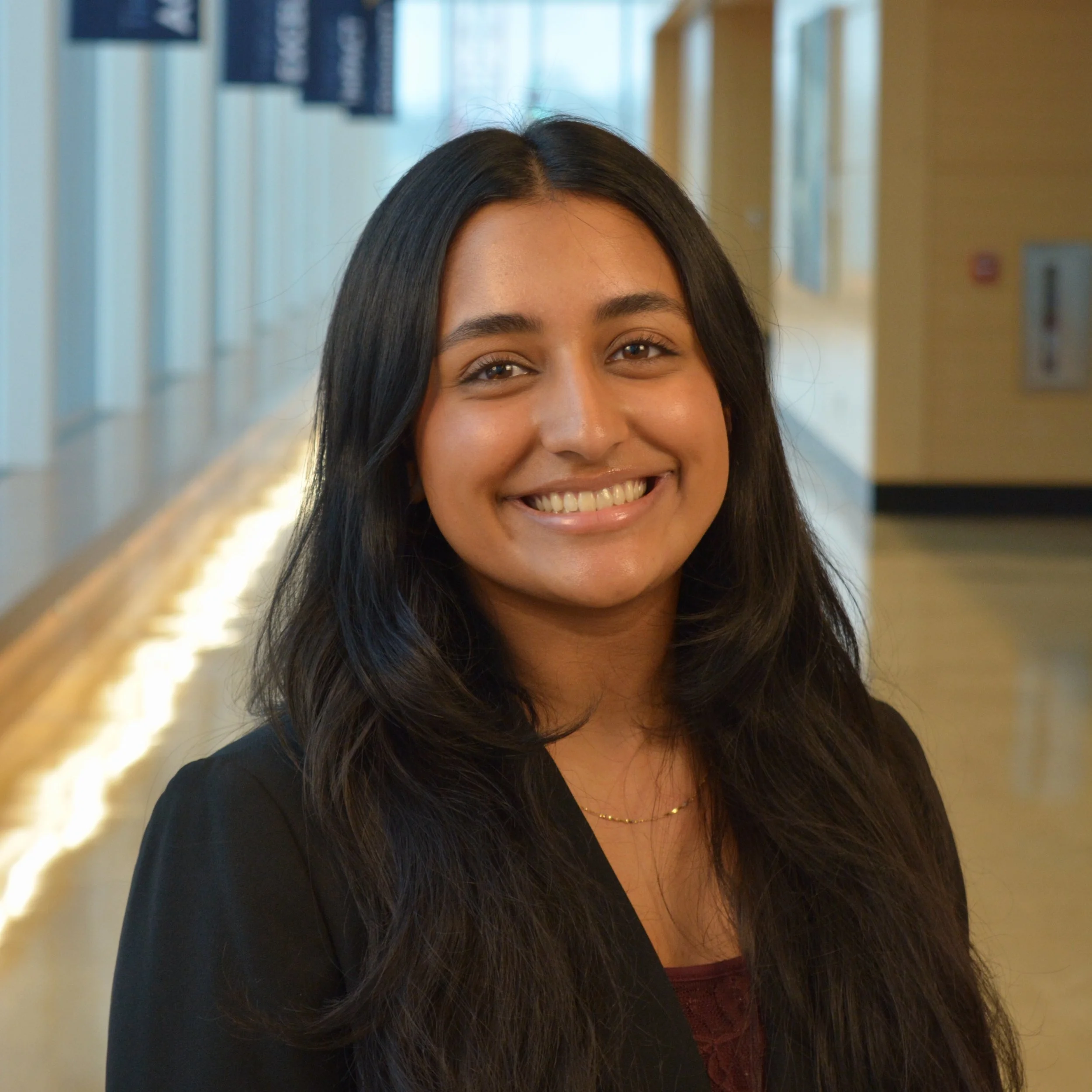 A young woman with long black hair and a bright smile standing in a well-lit indoor corridor.