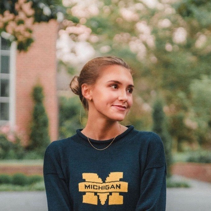 Young woman with her hair in a bun, wearing a dark blue Michigan sweatshirt and a delicate necklace, standing outdoors with a blurred natural and brick building background.