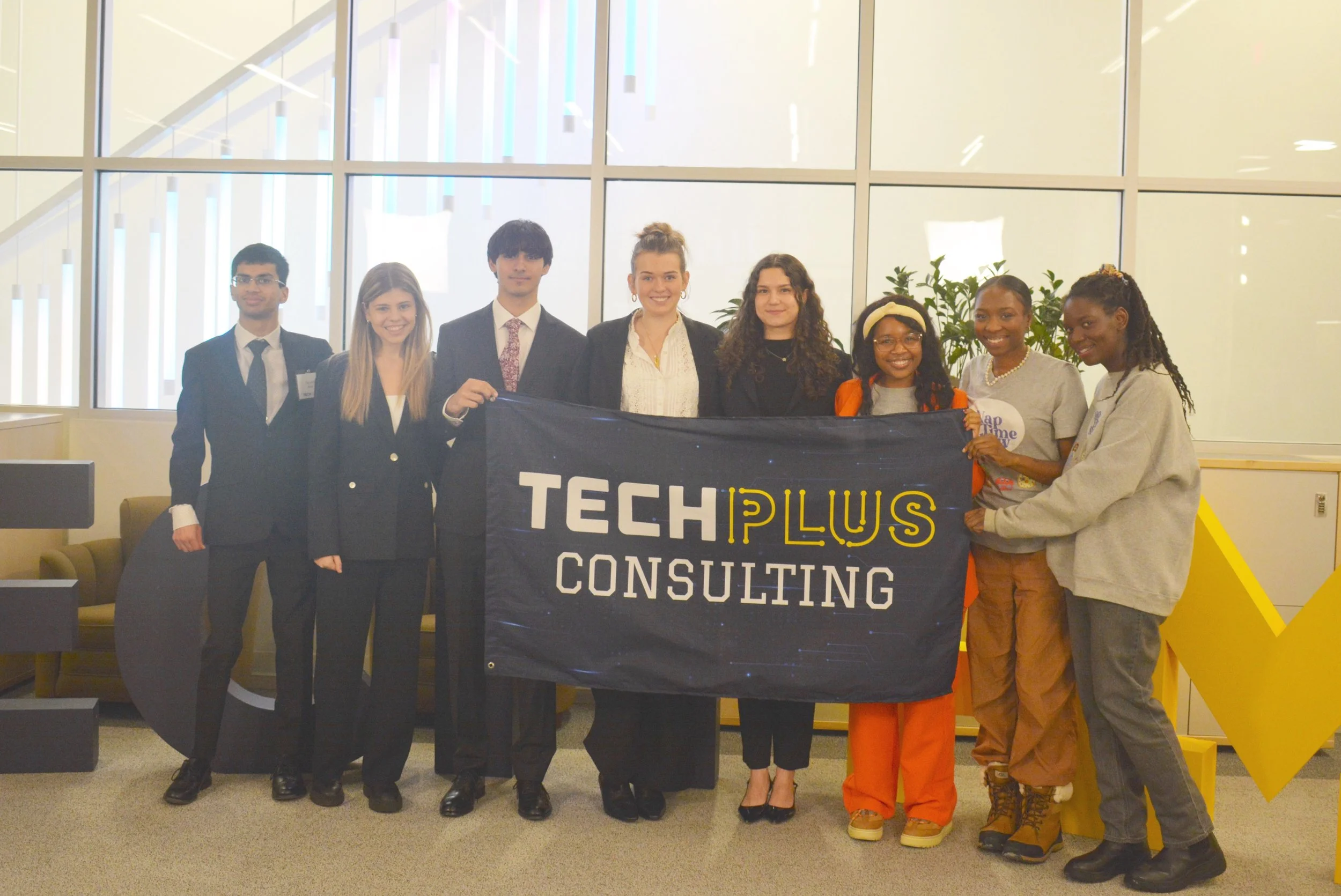 A diverse group of nine young professionals holding a banner that reads 'TECHPLUS CONSULTING' indoors in a modern office setting.