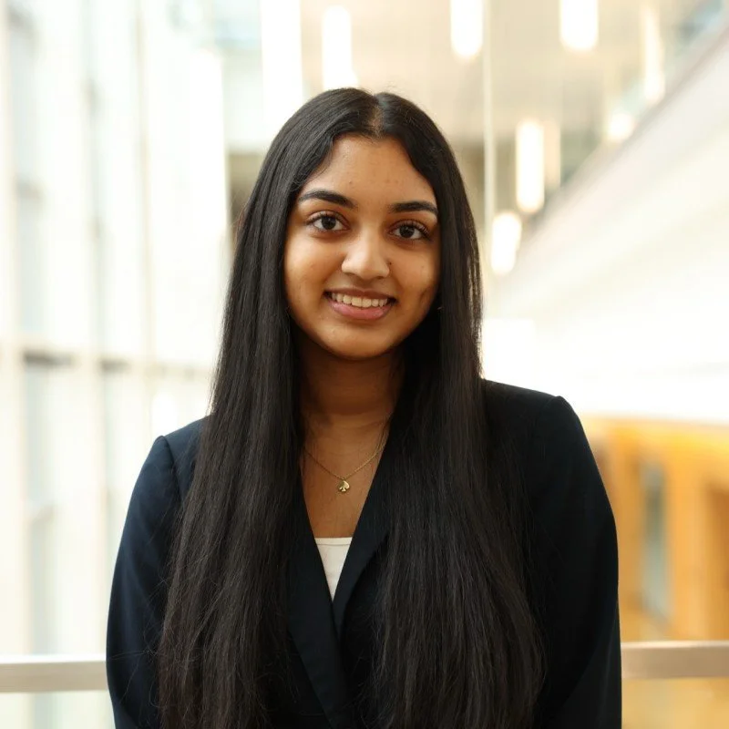 A young woman with long dark hair and a black blazer smiling indoors.