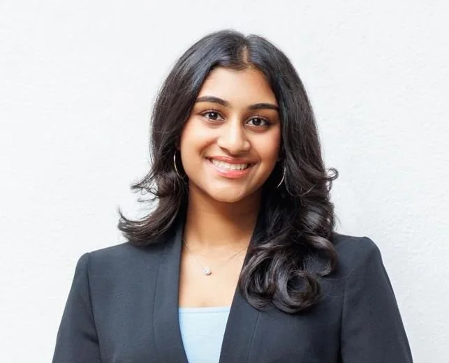 A young woman with dark hair smiling, wearing a black blazer and light-colored top, standing against a plain white background.