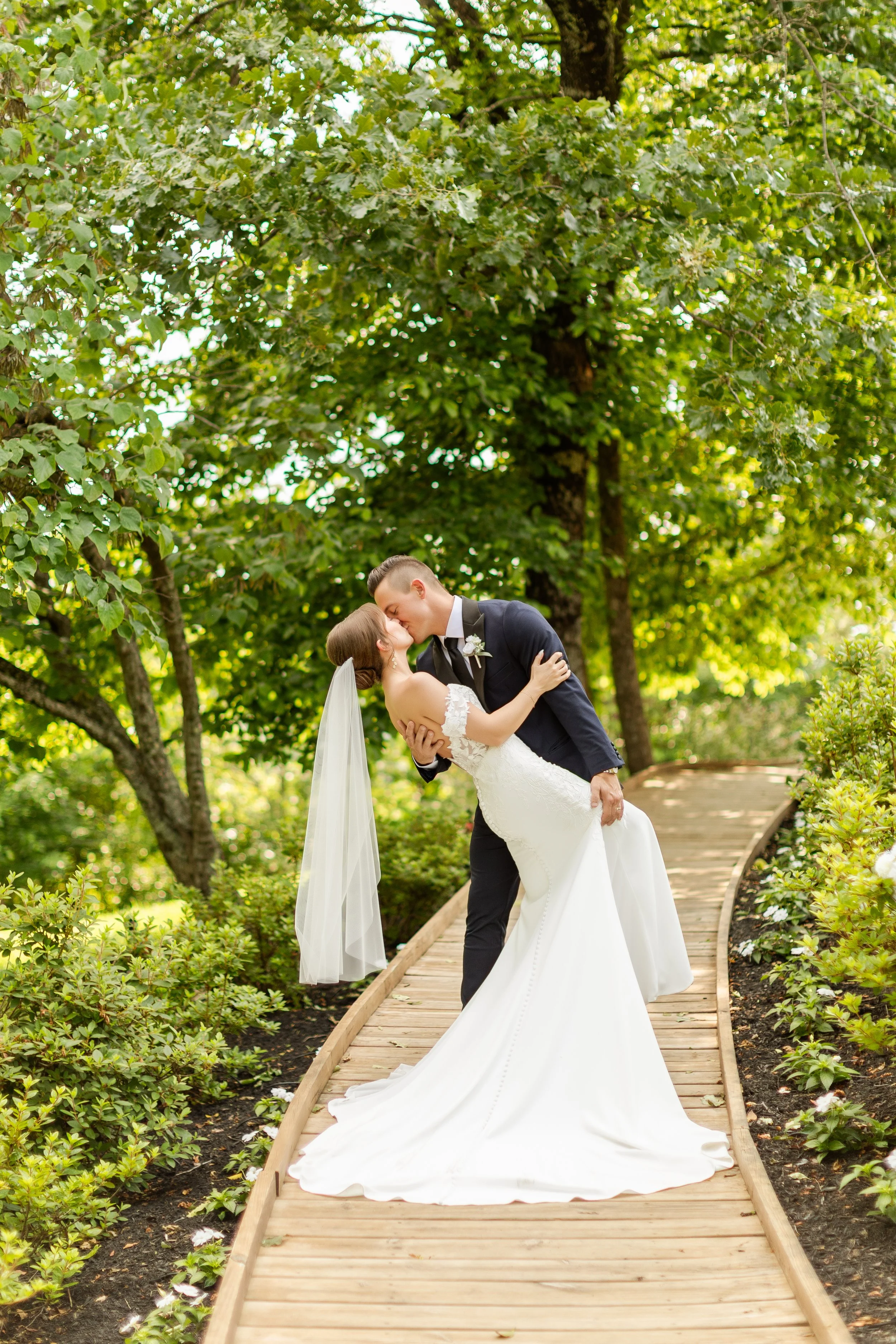 A bride and groom share a kiss on a wooden path surrounded by greenery, trees, and floral bushes in an outdoor setting.