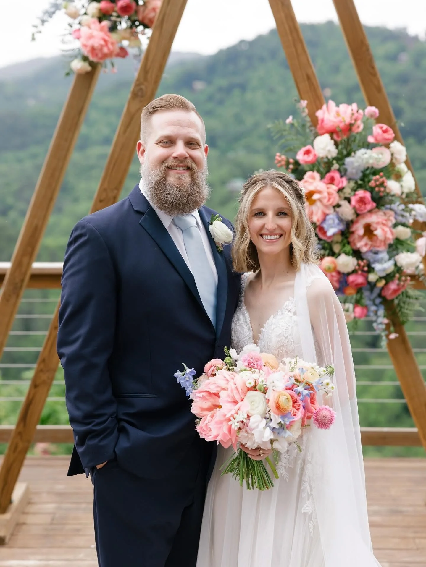A bride and groom at their wedding, standing together outdoors on a wooden platform with a lush green landscape in the background. The bride is holding a bouquet of pink, white, and purple flowers, and the wedding arch behind them is decorated with s
