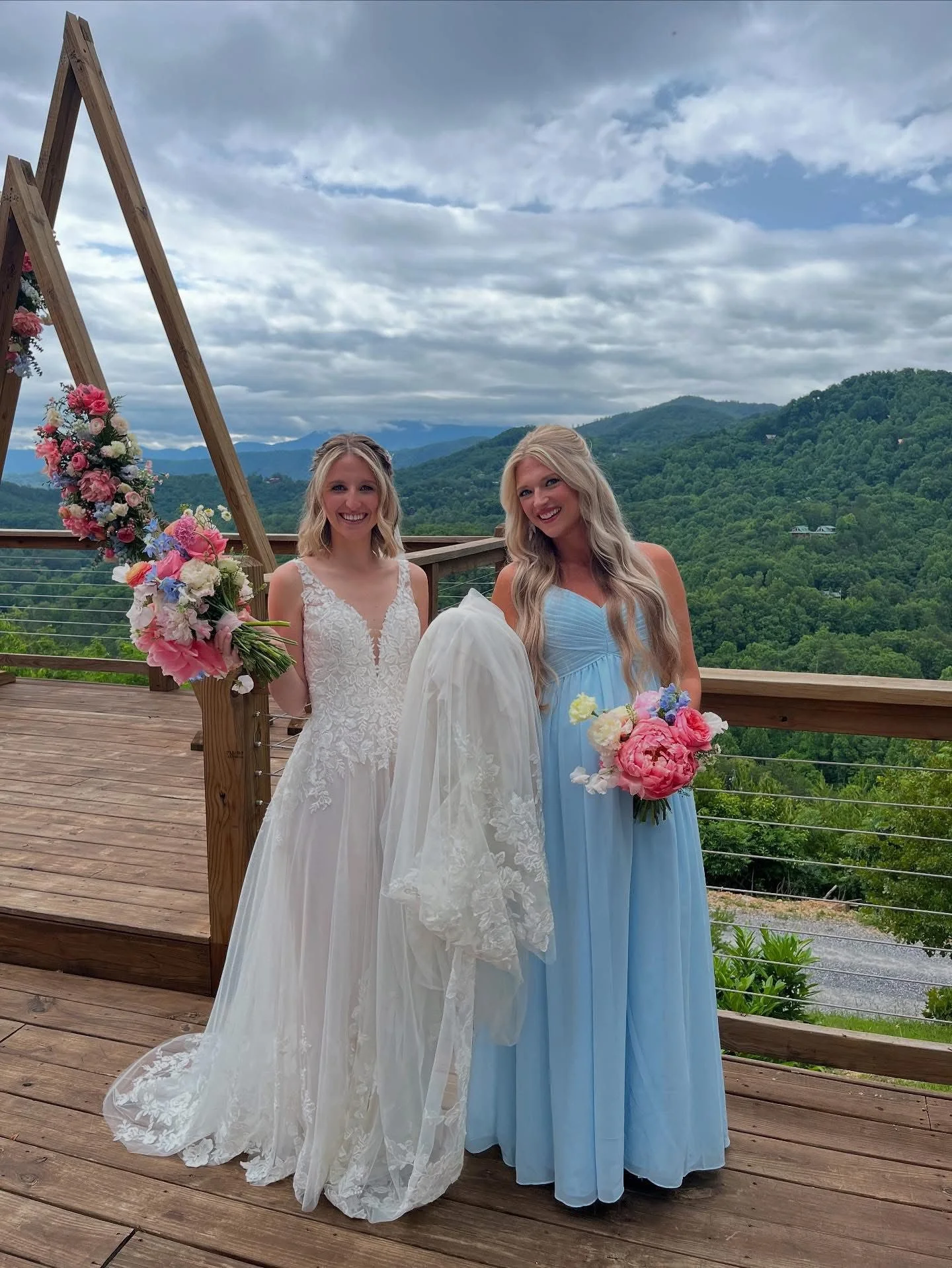 Two women in dresses holding bouquets, standing on a wooden deck outdoors with mountains and cloudy sky in the background.