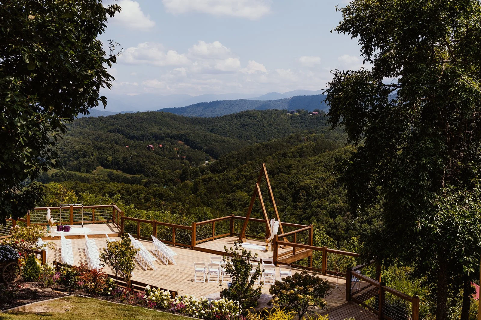 Outdoor wedding ceremony setup on a wooden deck with white chairs and an easel, overlooking a lush green mountain landscape under a partly cloudy sky.