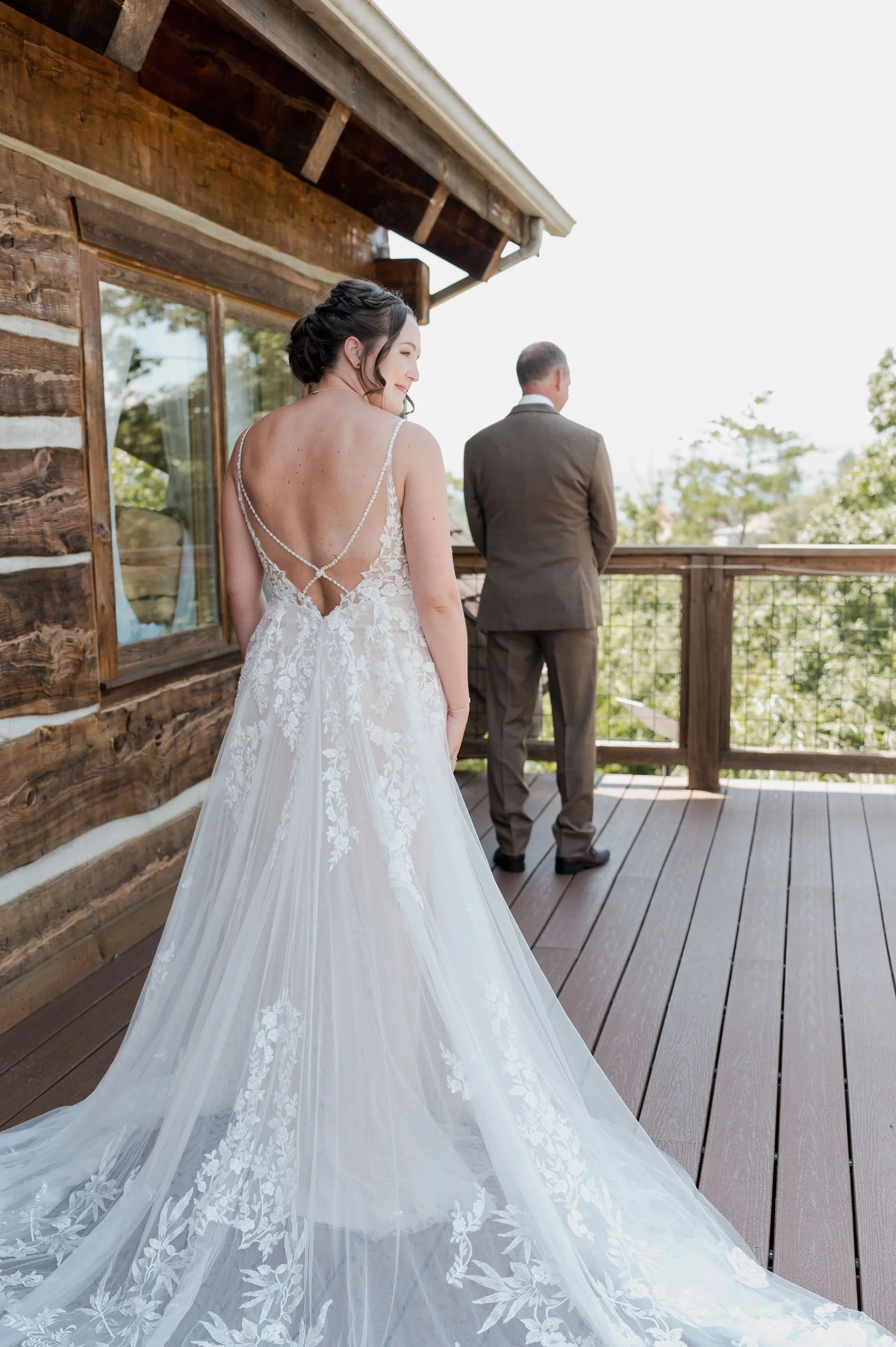 A bride in a white wedding gown with lace details and an open back, standing on a wooden balcony with a groom in a brown suit in the background, outside of a rustic wooden house.