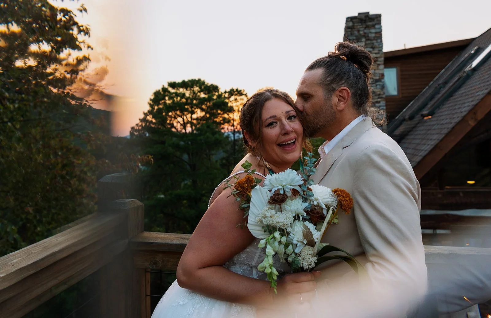 A bride and groom sharing a tender moment outdoors during sunset, with the groom kissing the bride on her forehead while she holds a bouquet of white and brown flowers.